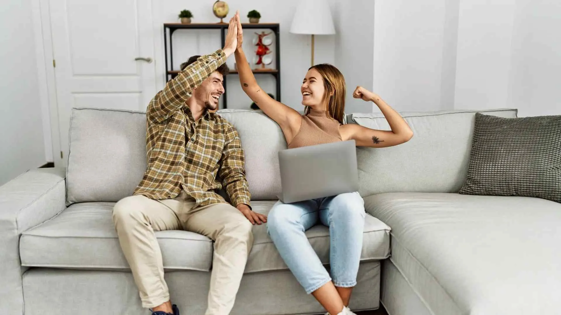 Young man and woman sitting on couch giving high five with laptop, celebrating success or achievement at home