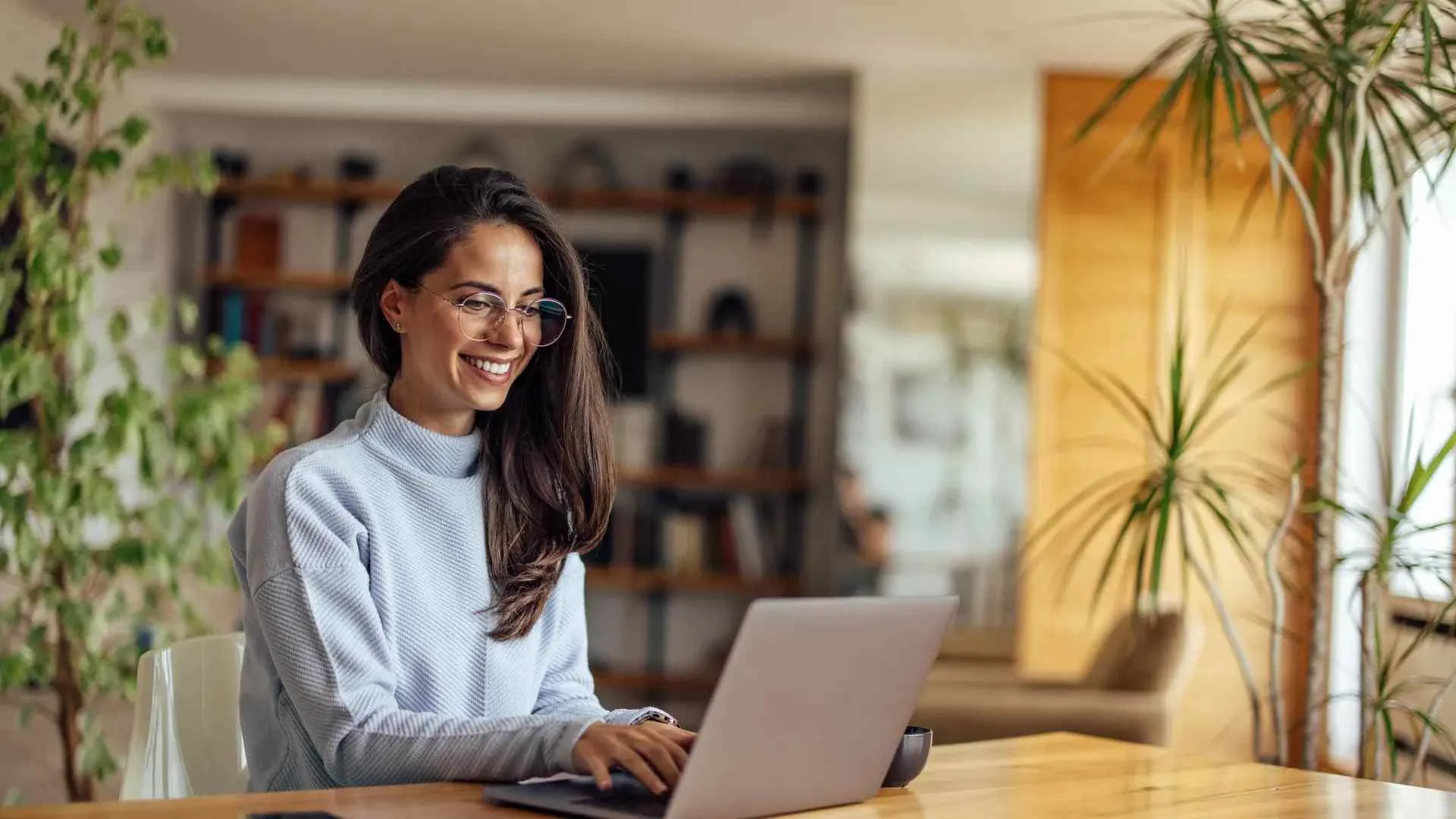 Smiling woman with glasses working on laptop at wooden table in bright home office surrounded by plants.