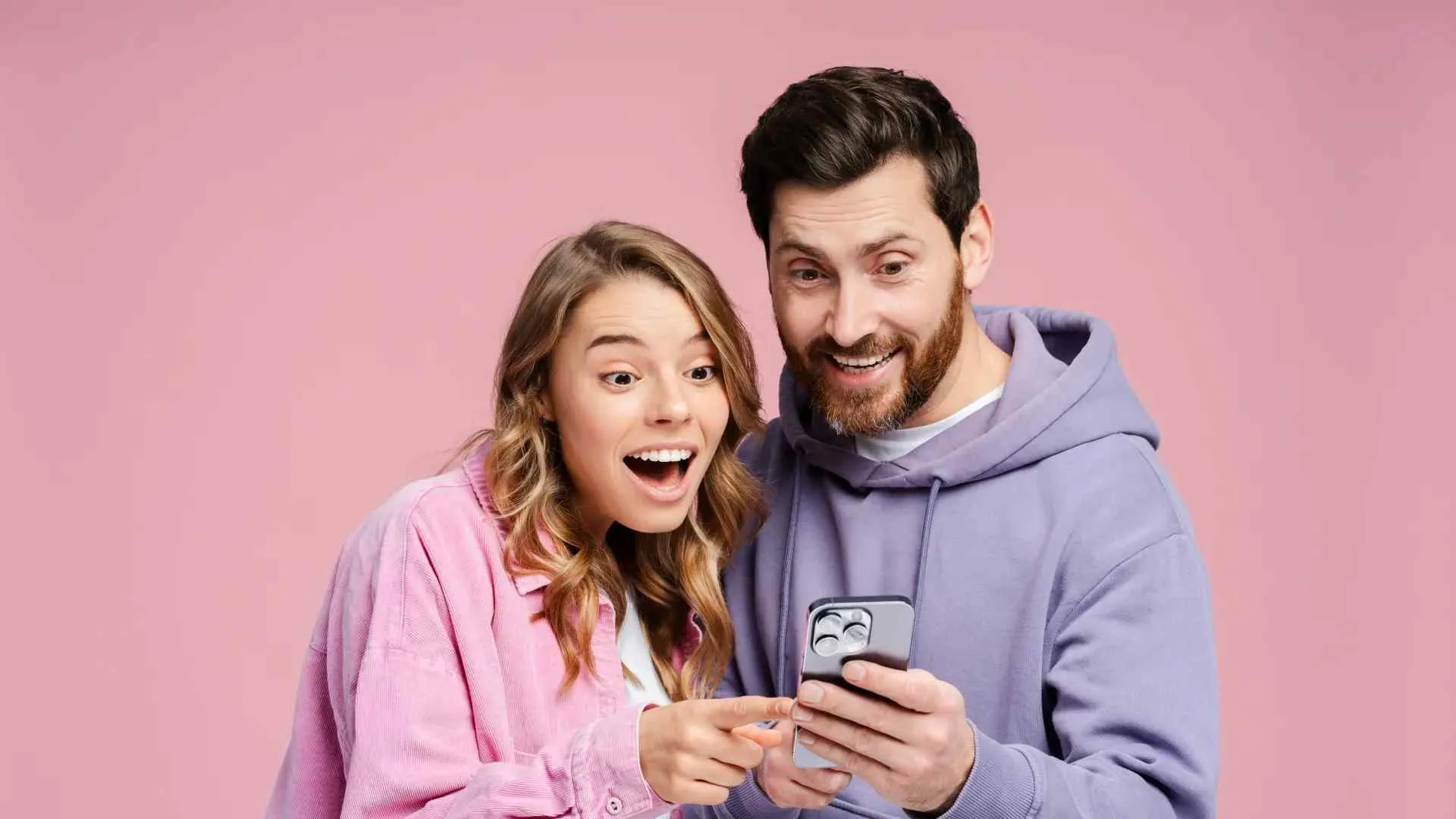 Excited young couple looking at smartphone screen together against a pink background.