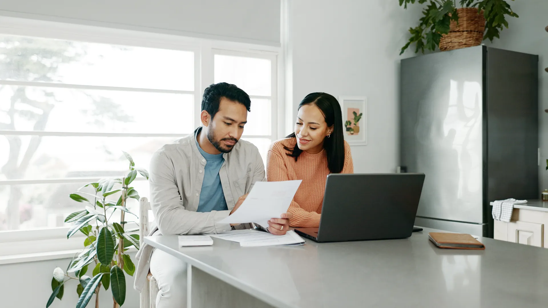 Couple reviewing documents together at kitchen table with laptop and indoor plants in natural light.