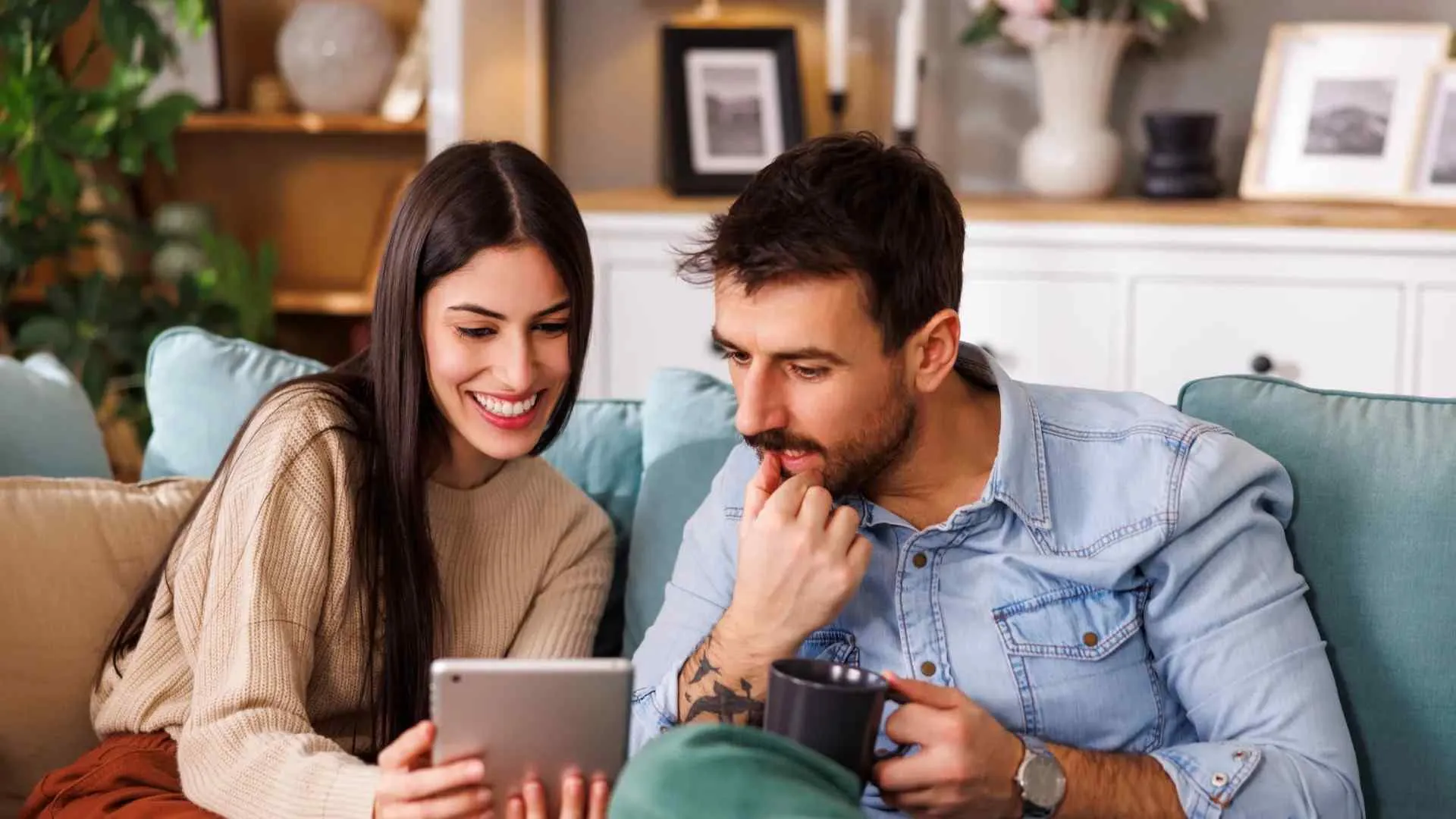 Smiling young couple sitting on couch looking at tablet together in cozy living room.