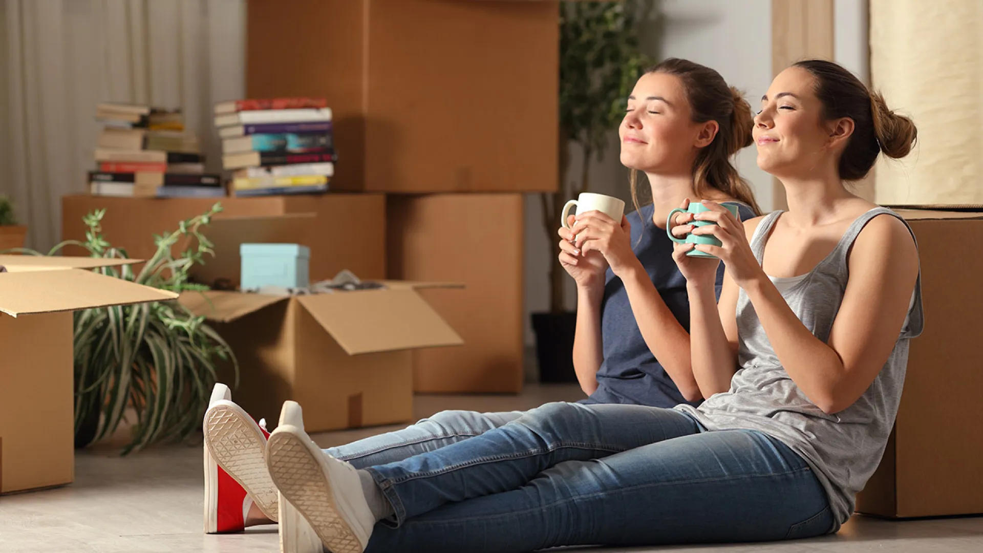 Two young women relaxing with coffee while sitting on floor surrounded by moving boxes and stacked books.