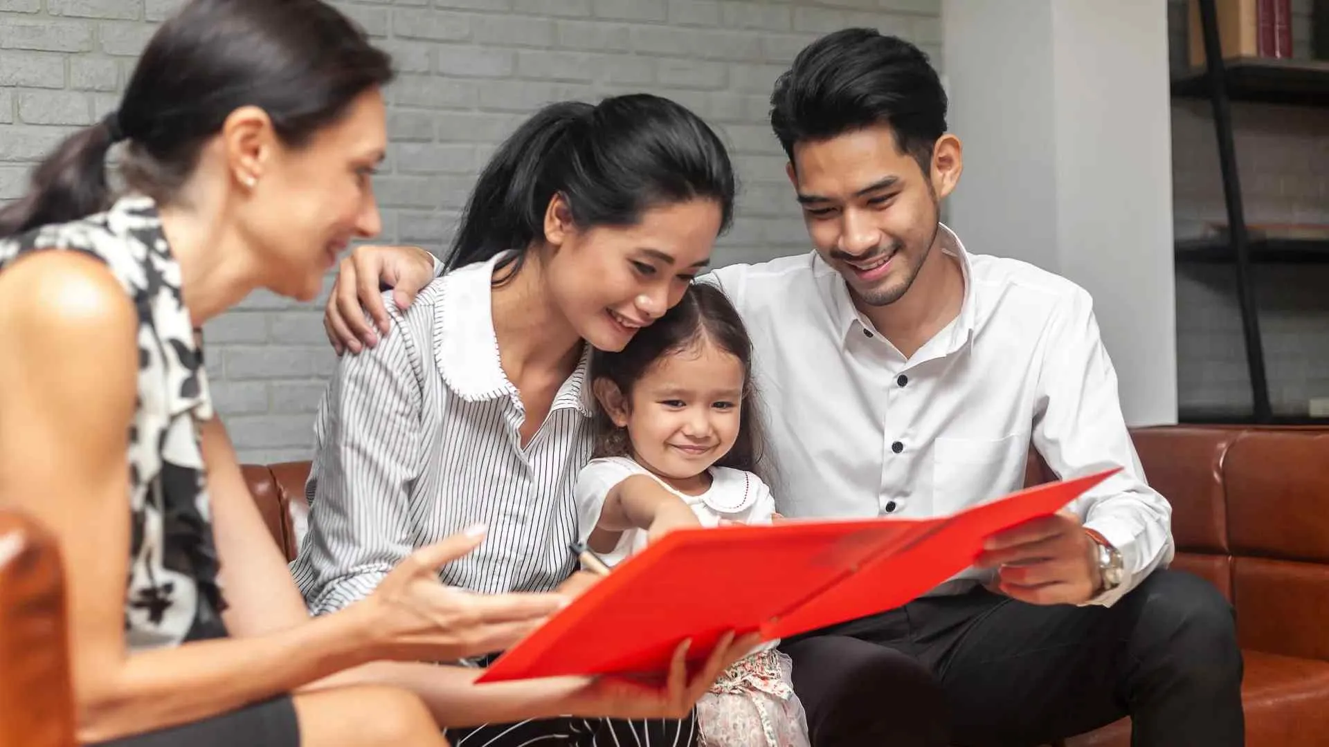 Family with young daughter smiling while looking at a red photo album in a cozy living room.