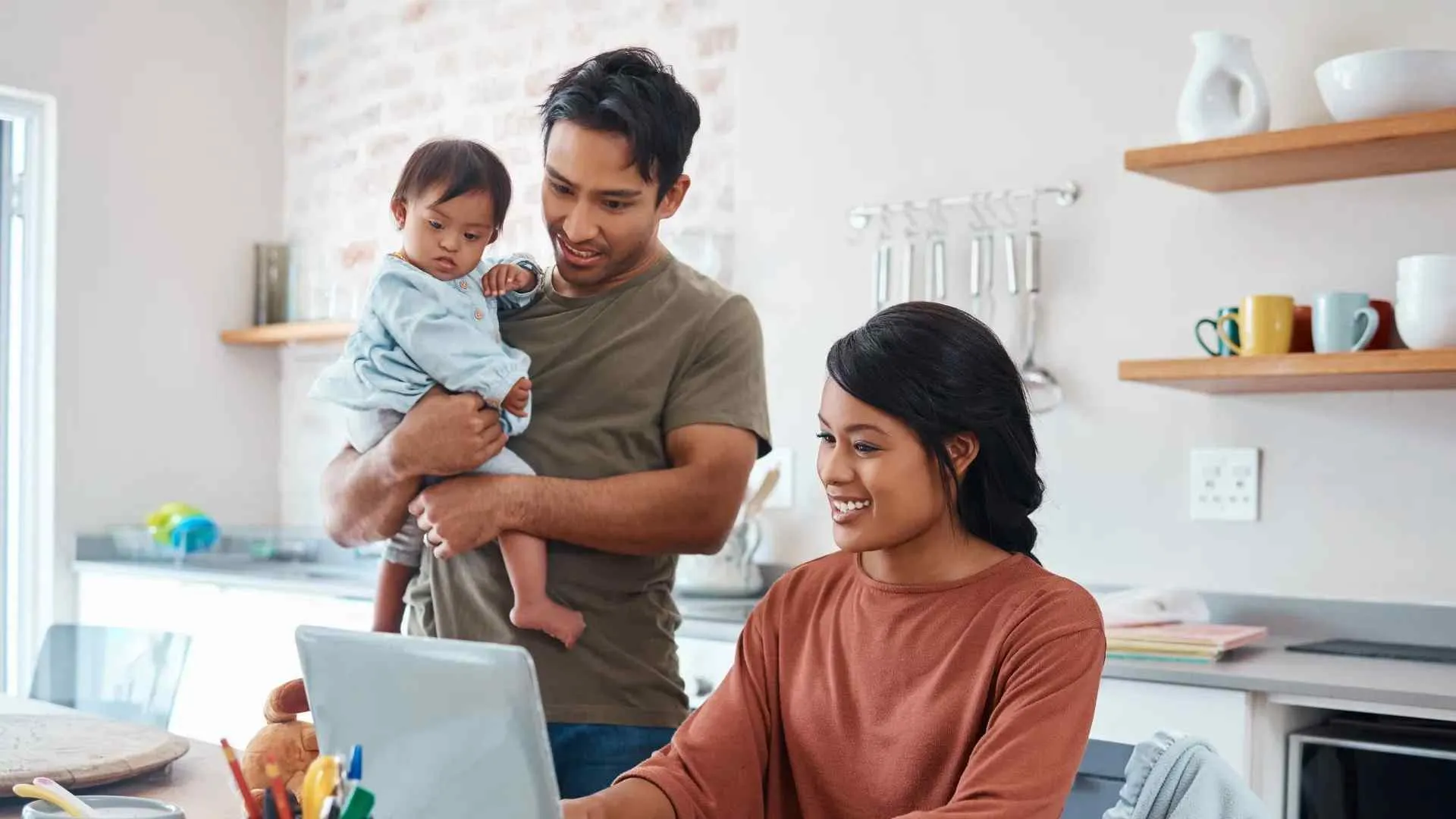 Smiling young parents with toddler using laptop together in modern kitchen at home