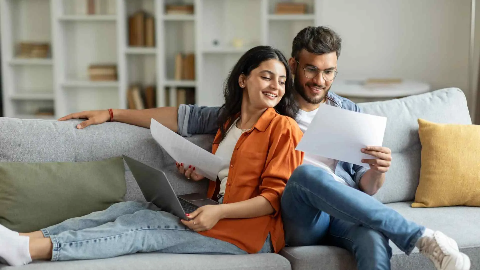 Young couple sitting on sofa reviewing documents with laptop, smiling and relaxed in bright living room.