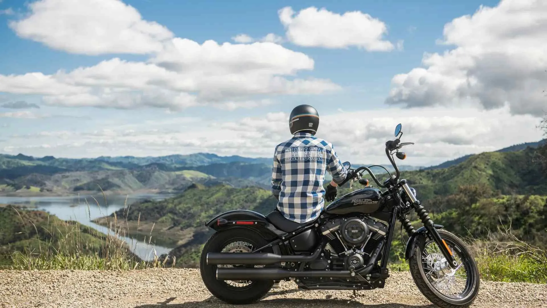 Man in checkered shirt sitting on black motorcycle overlooking scenic river valley under cloudy sky.