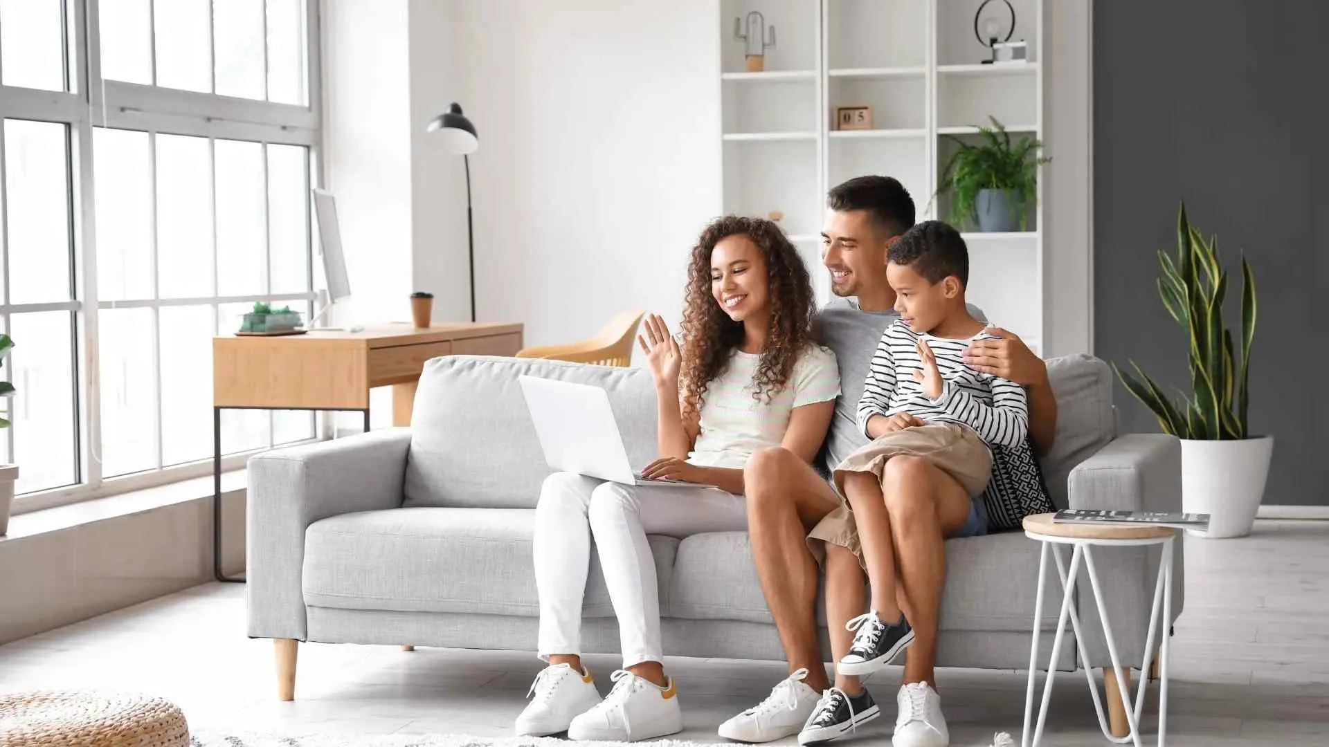 Smiling family of three sitting on a couch video calling on a laptop in a bright living room