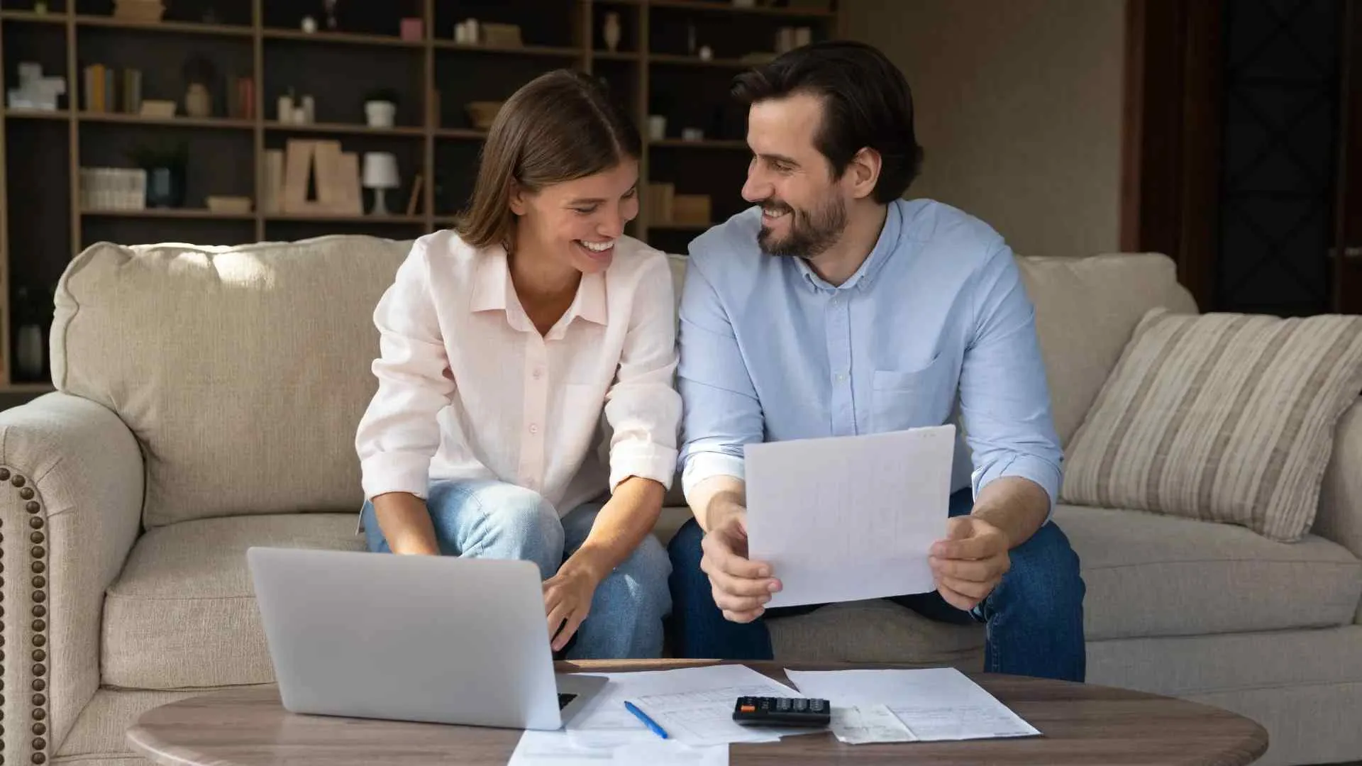 Couple sitting on sofa reviewing documents and using laptop at a coffee table with papers and calculator