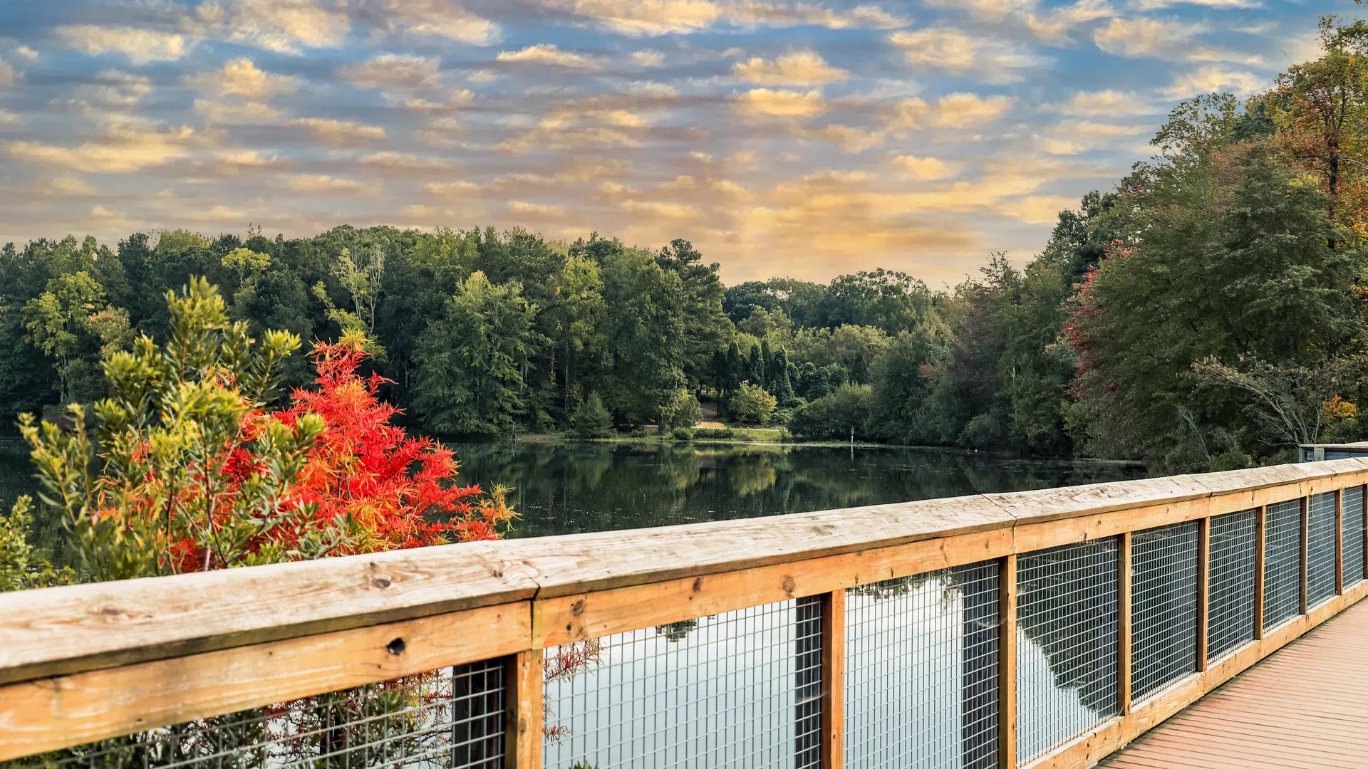 Wooden railing along a peaceful lake with colorful autumn trees and a cloudy sky during sunset.