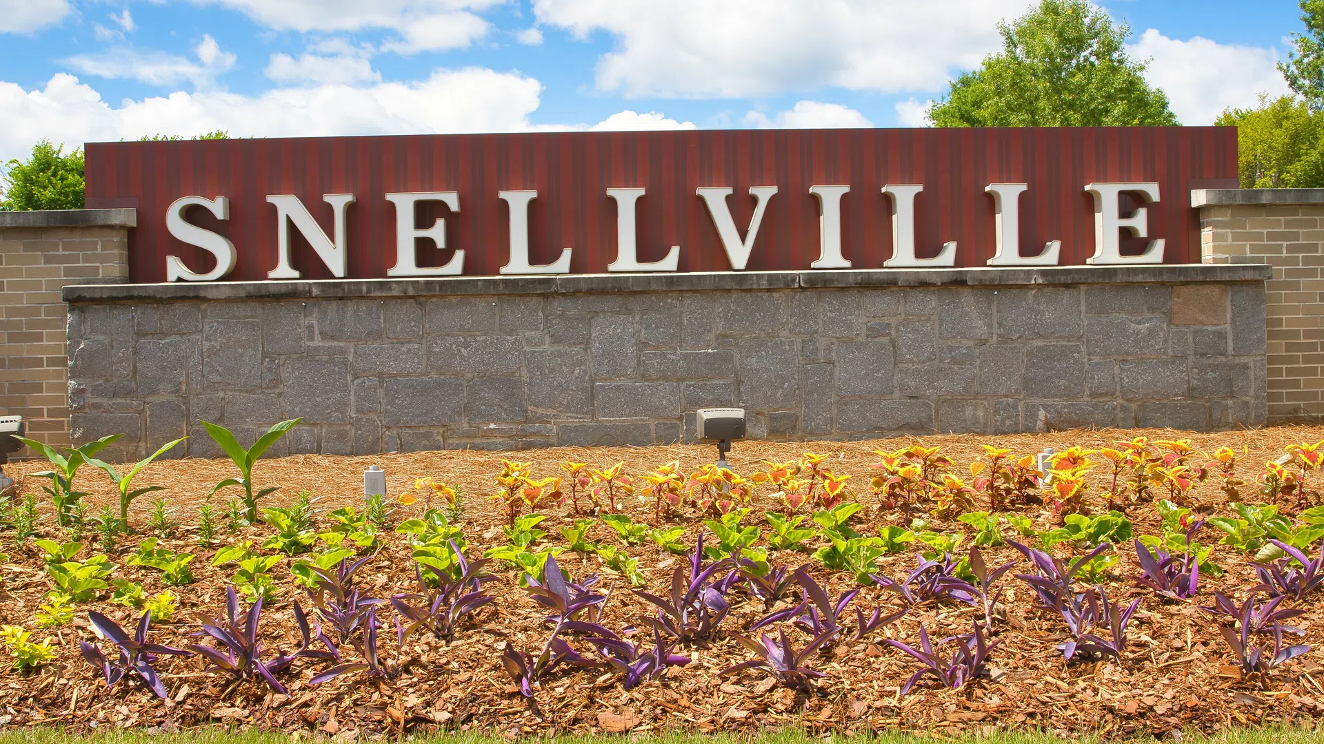 Entrance sign for Snellville with stone wall and landscaped garden under a bright blue sky.