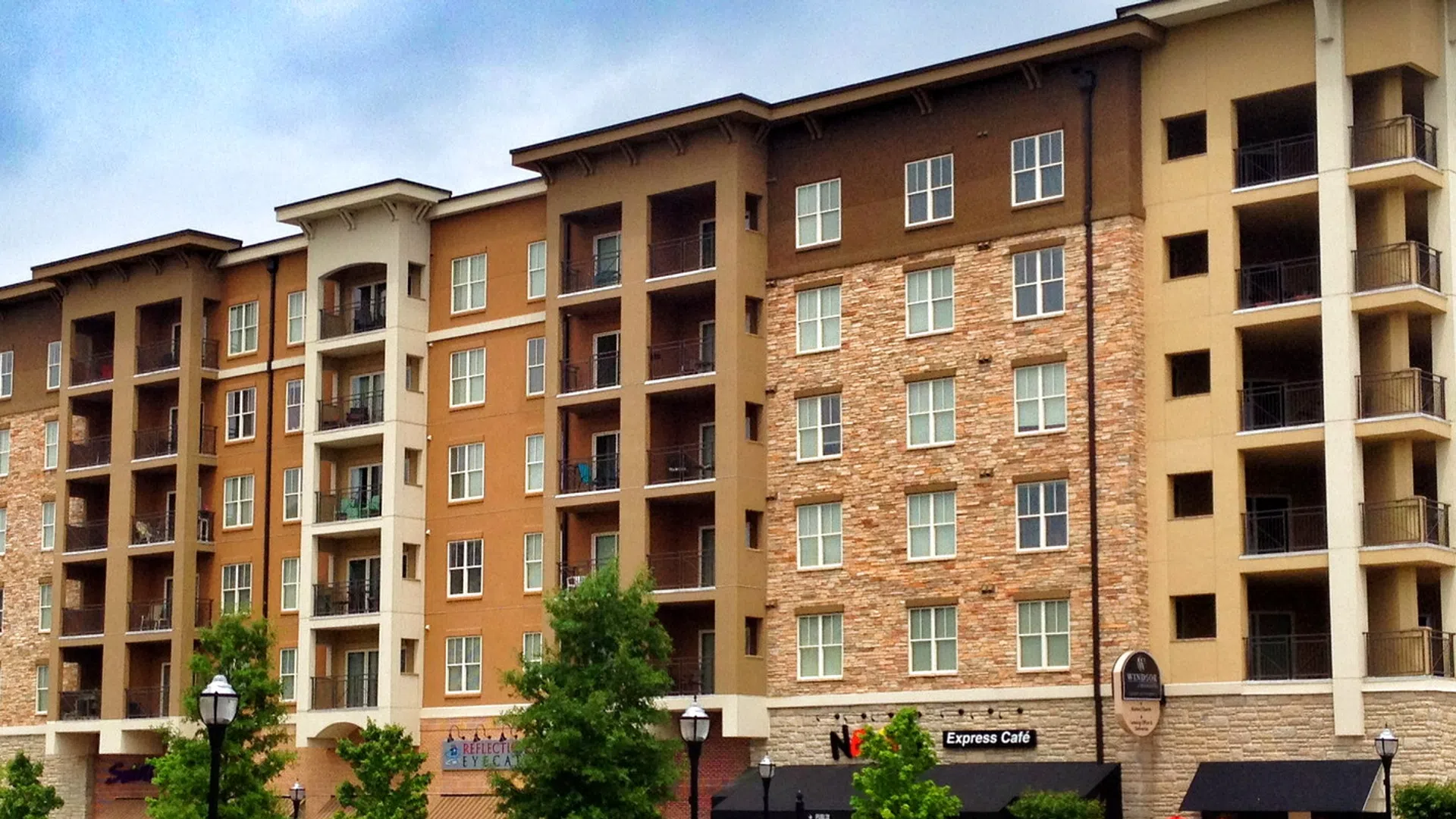 Modern multi-story apartment building with balconies, stone and stucco facade, and street lamps in a residential area.