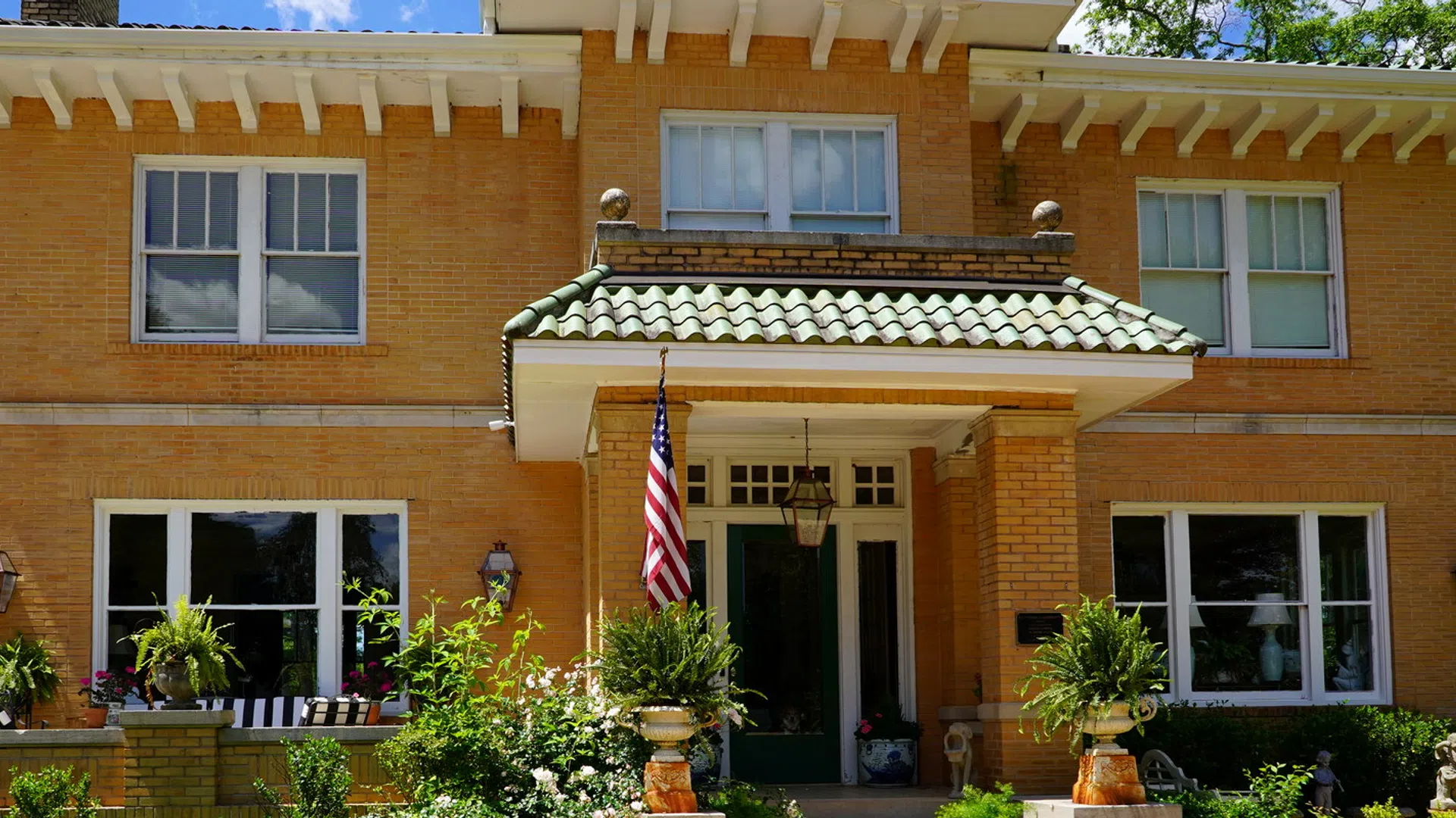 Two-story yellow brick house with green tile roof, front porch, American flag, and lush potted plants in sunny weather