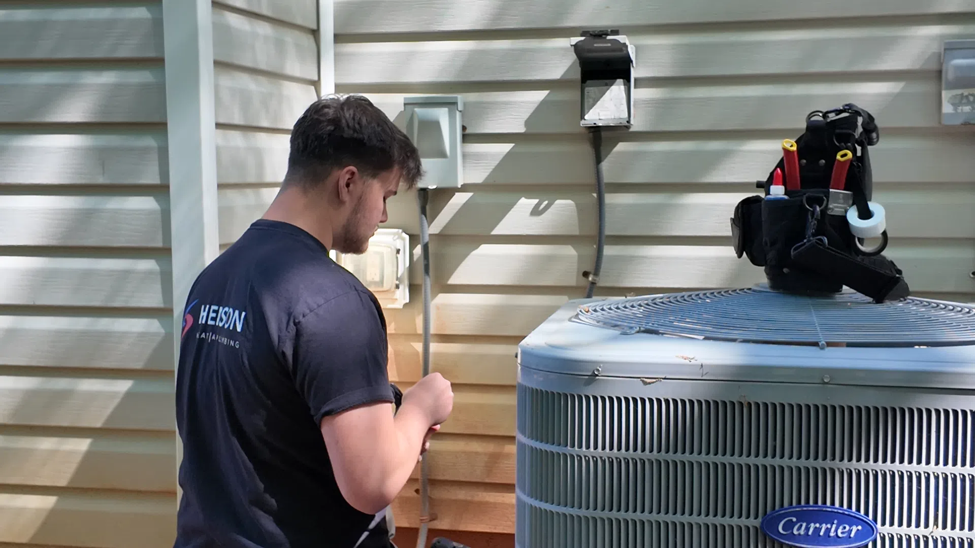 Technician kneeling beside a Carrier air conditioning unit outside a house during repair work.