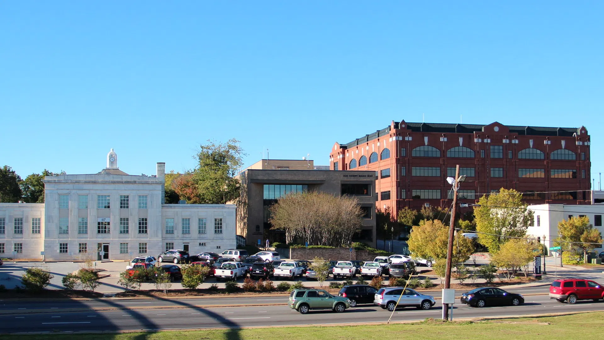 Urban street scene with historic white building, modern red brick building, cars parked and trees on a sunny day.