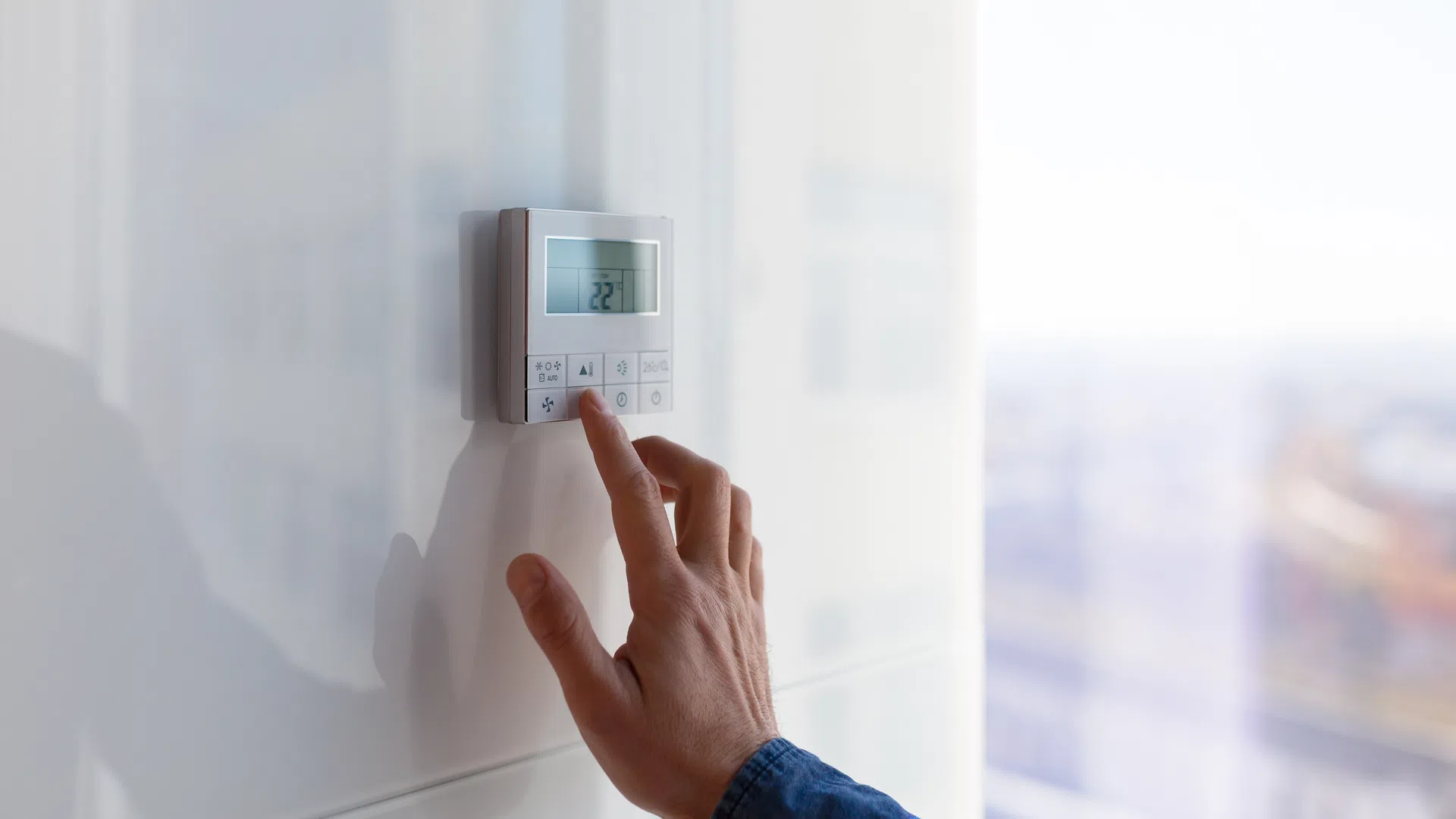 Hand adjusting modern digital thermostat on white wall with visible temperature display.