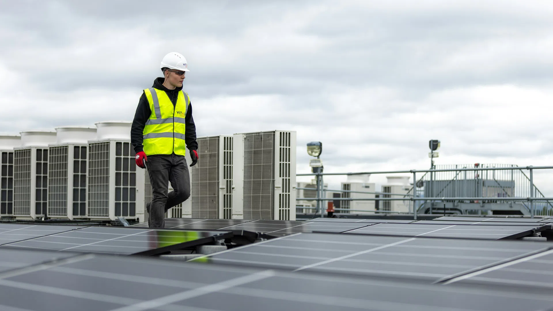 Technician in safety gear inspects solar panels on rooftop with industrial HVAC units in the background under a cloudy sky