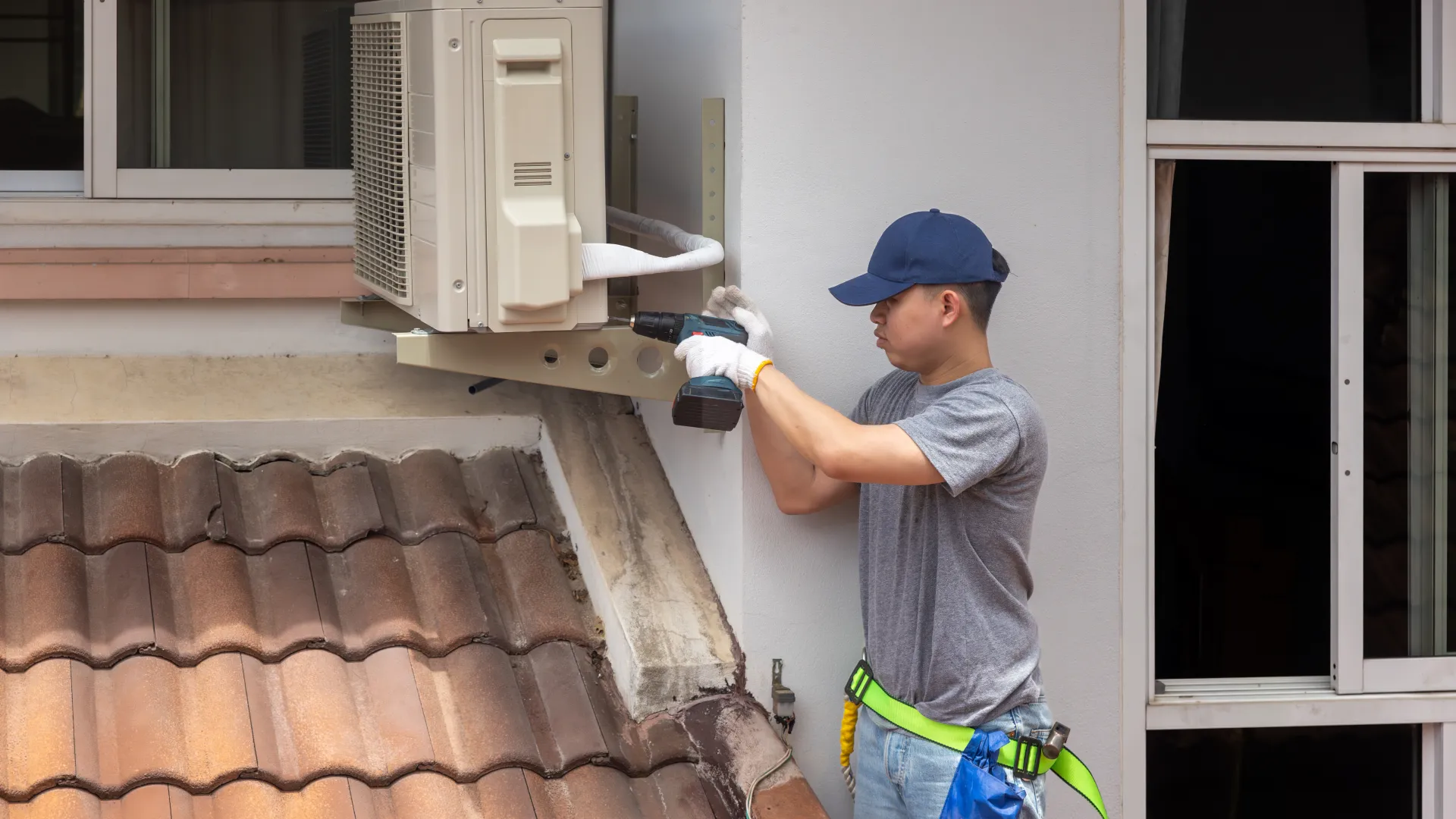 Technician wearing gloves and cap installs an air conditioning unit on a house exterior wall above a tiled roof.