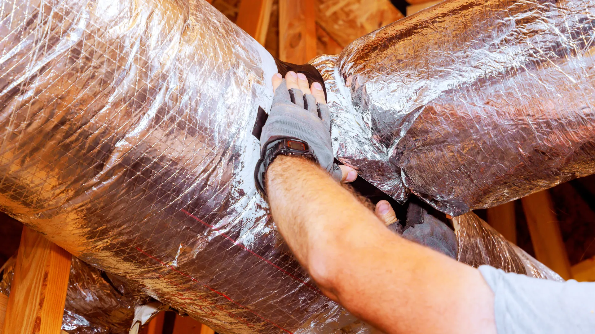 Technician wearing gloves working on insulated HVAC ducting in a building attic with wooden beams.
