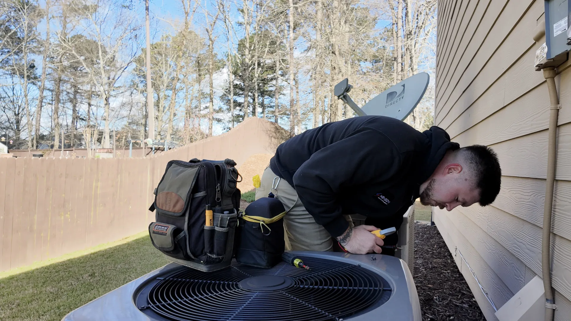 Technician inspecting a Rheem HVAC unit outside a house on a clear day with tool bag on the unit
