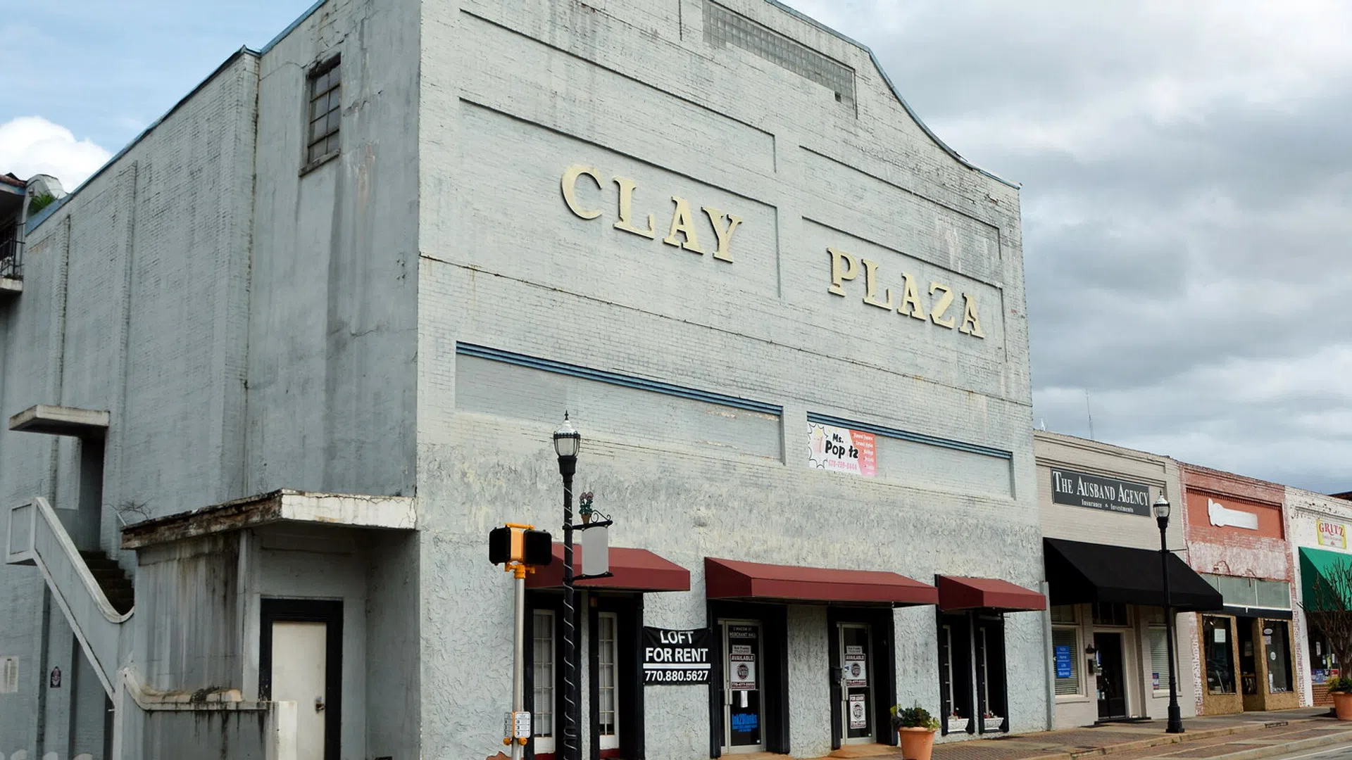 Clay Plaza building with gray facade and red awnings in a small downtown shopping area under cloudy sky.