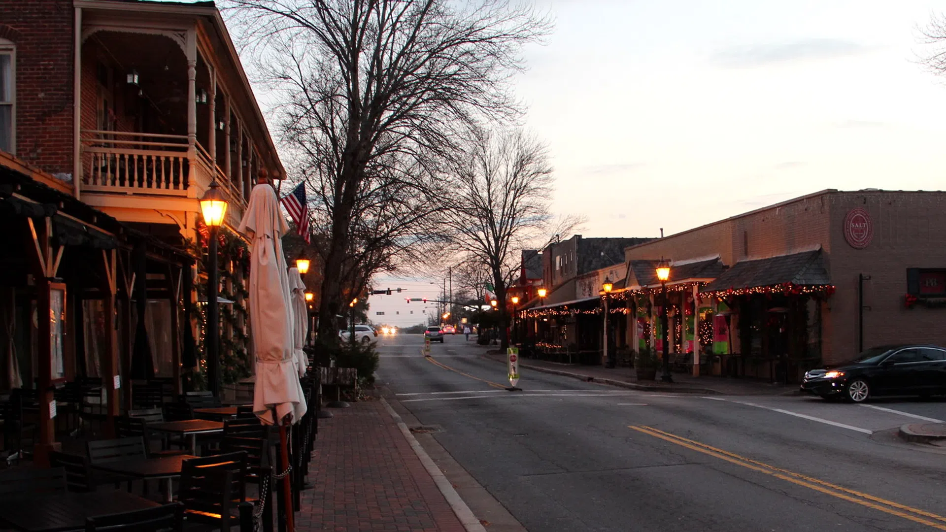 Empty street scene at dusk with historic buildings, street lamps, and outdoor seating on a small town main street.