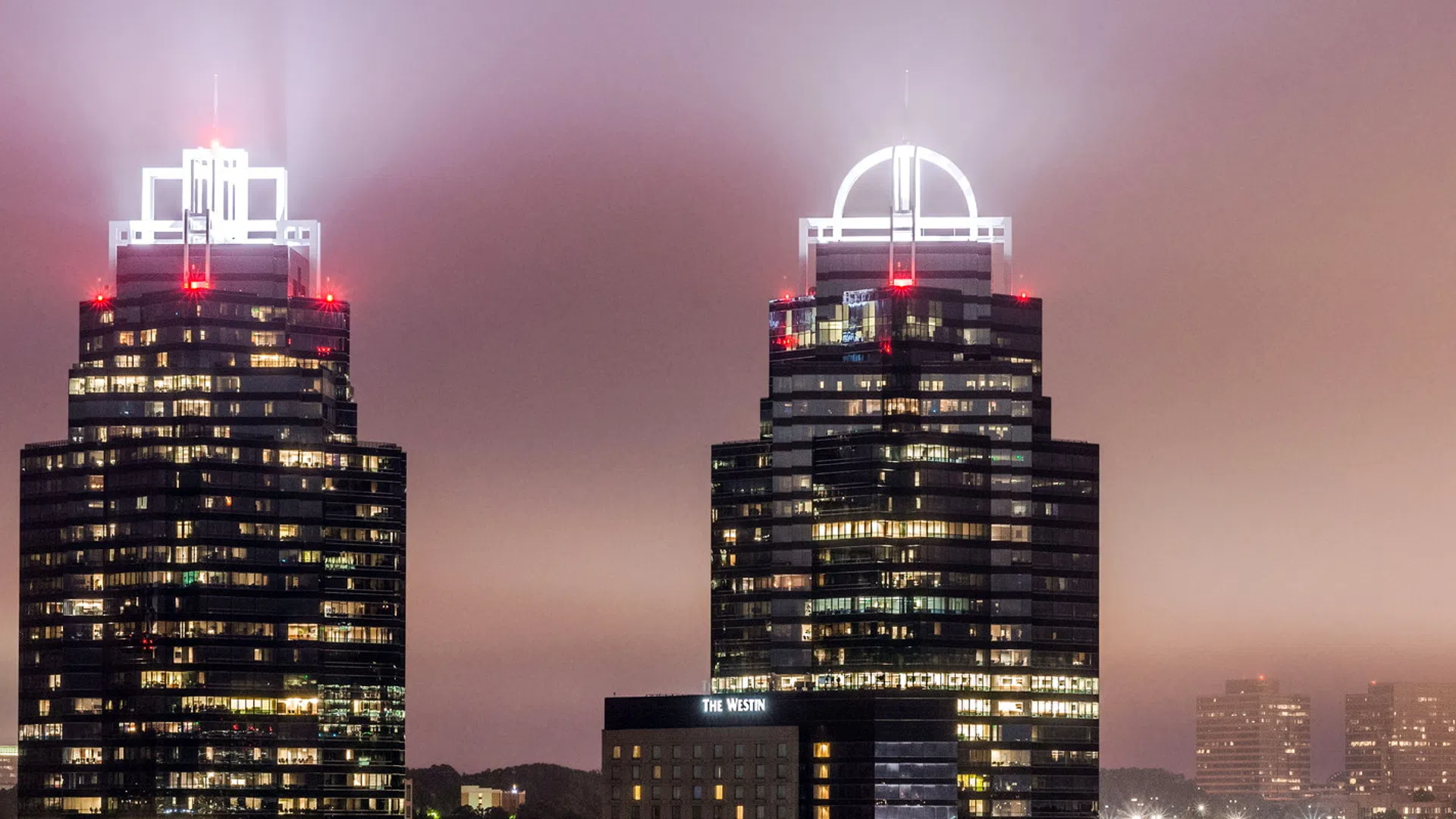 Twin illuminated skyscrapers with distinctive crowns glowing through evening fog in an urban cityscape at dusk
