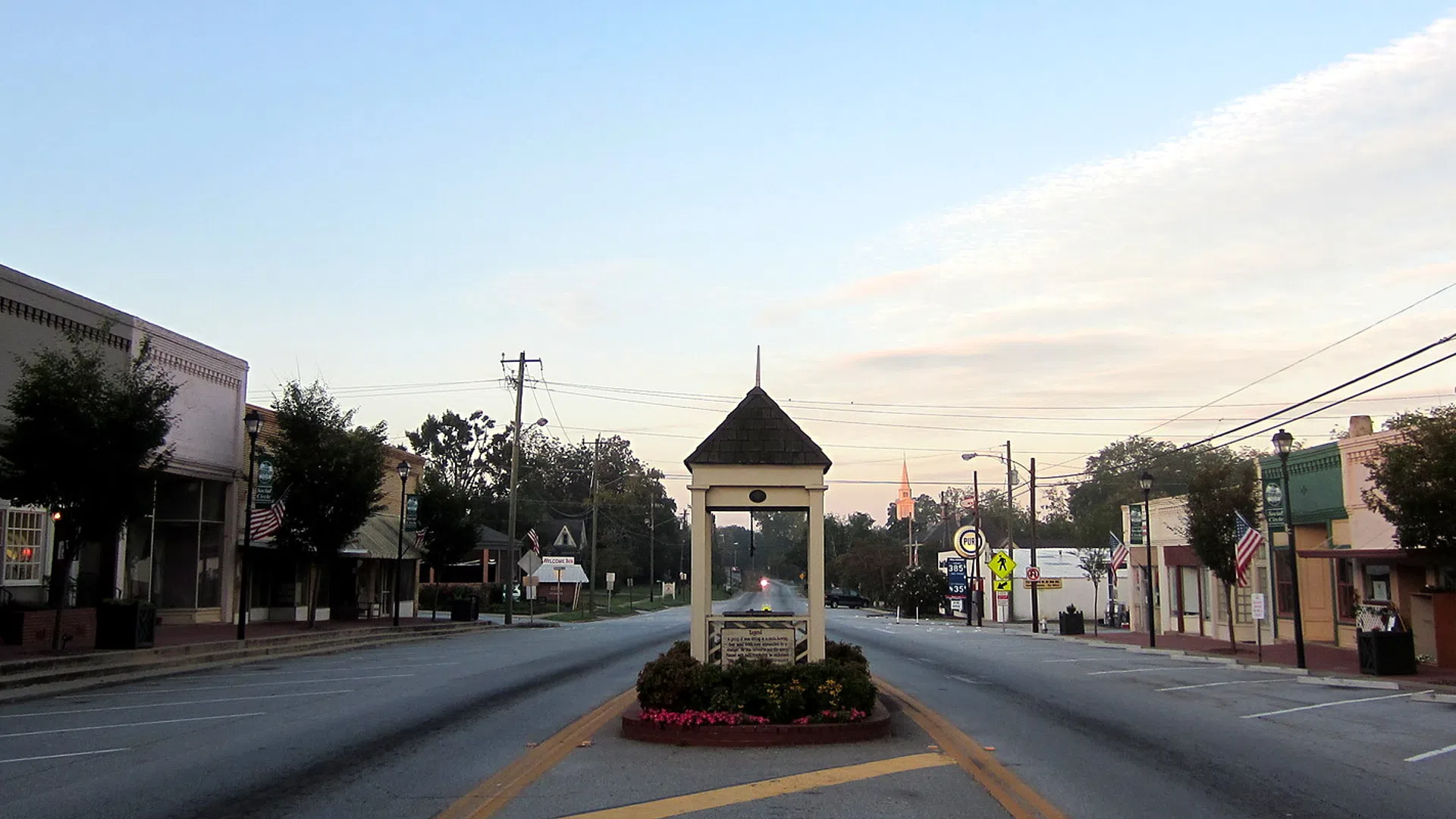 Small town main street at sunset with a monument in the center median and historic buildings lining both sides.
