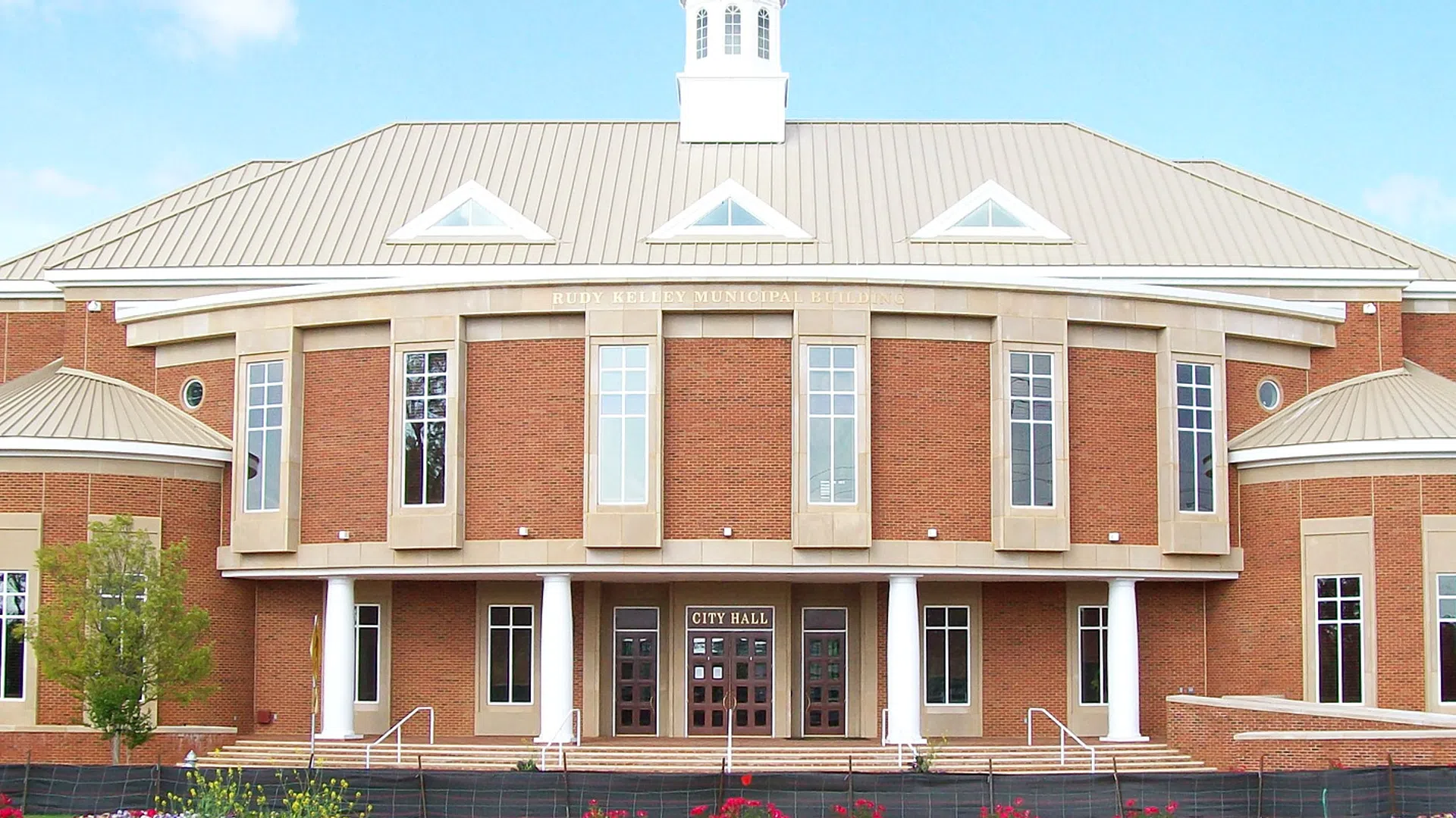 Red brick city hall building with large windows and a central entrance under a cloudy blue sky.