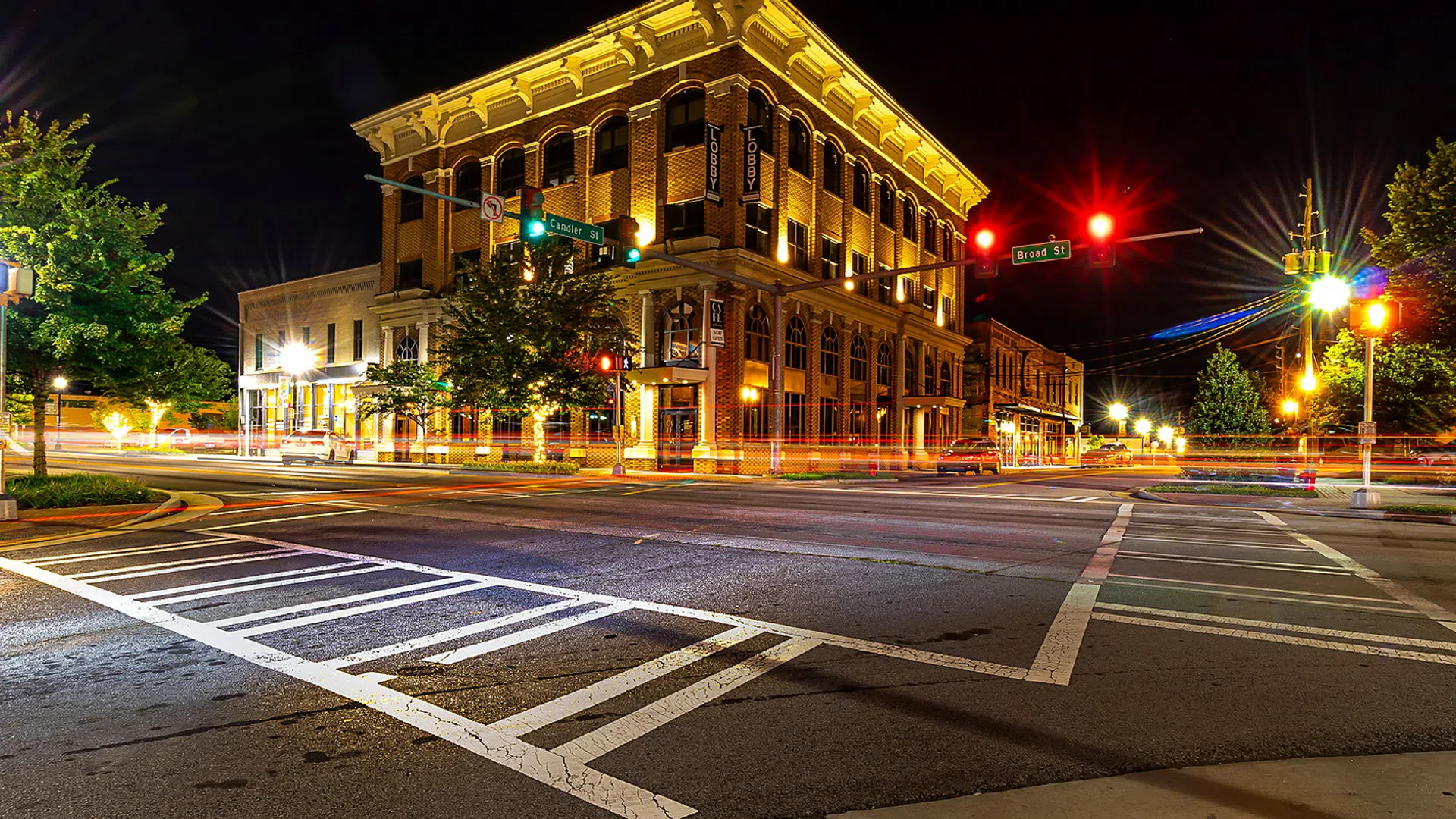 Nighttime street intersection with historic lit building, traffic lights, and street trees under a dark sky.
