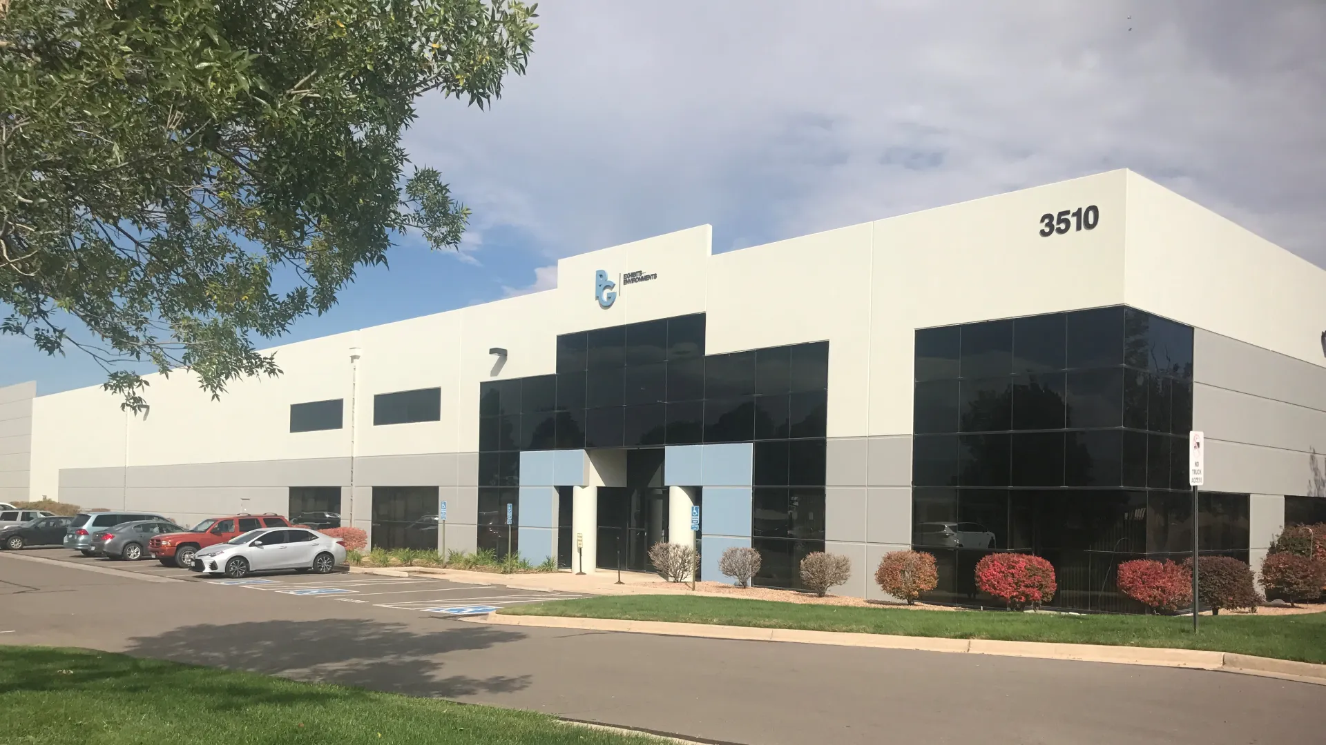 Modern industrial building with reflective glass windows and parked cars under a partly cloudy sky