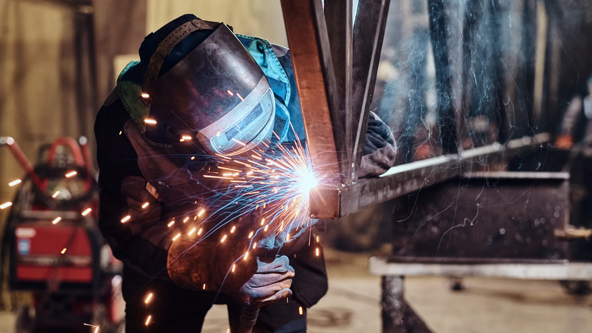 Welder wearing protective gear sparks flying while welding metal structure in industrial workshop