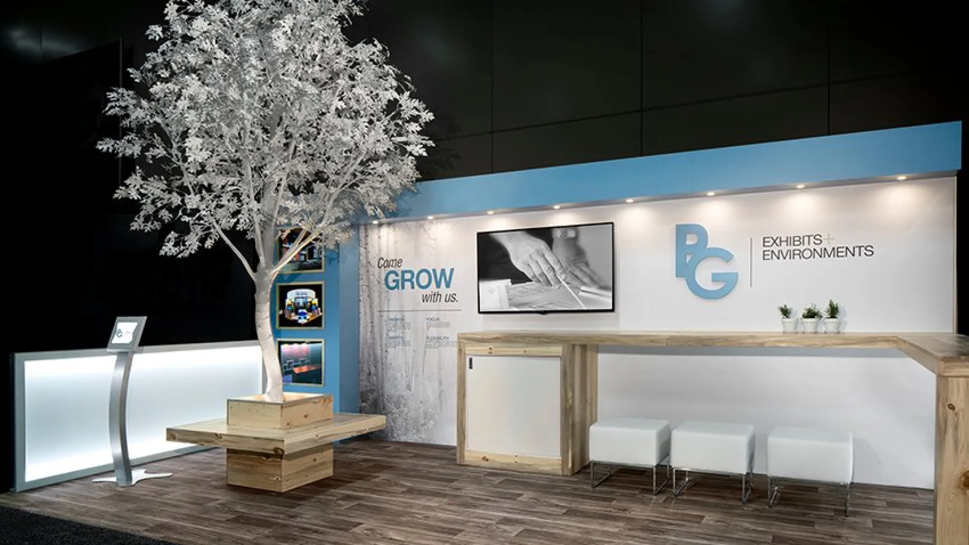 Modern trade show booth with a white tree, wooden counters, stools, and PG Exhibits branding on a white and blue backdrop