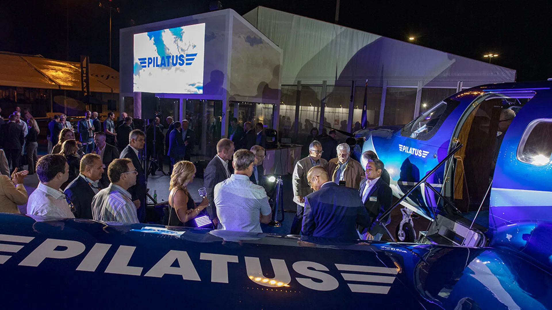Crowd of people gathered around a blue Pilatus aircraft at night with a large Pilatus logo displayed on screen.