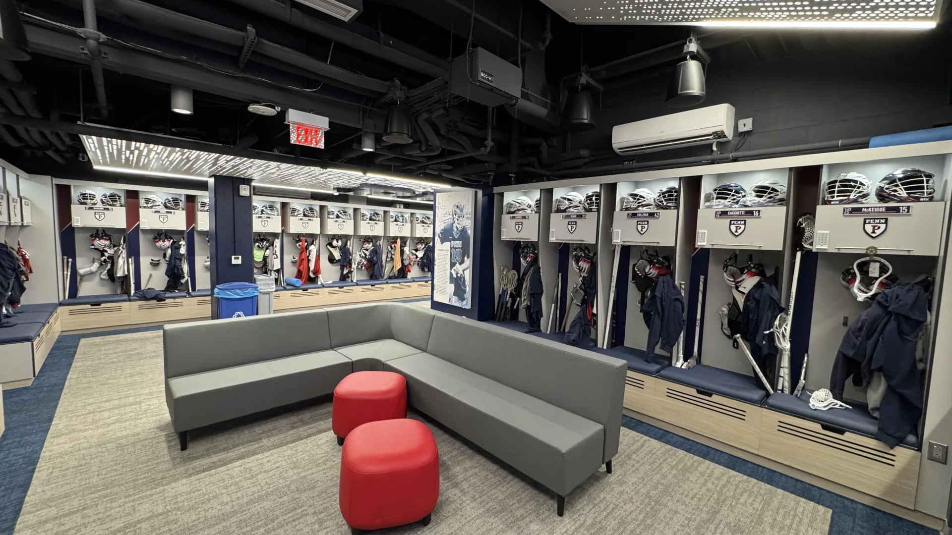 Modern sports team locker room with gray seating, red ottomans, hockey gear, and illuminated ceiling panels.