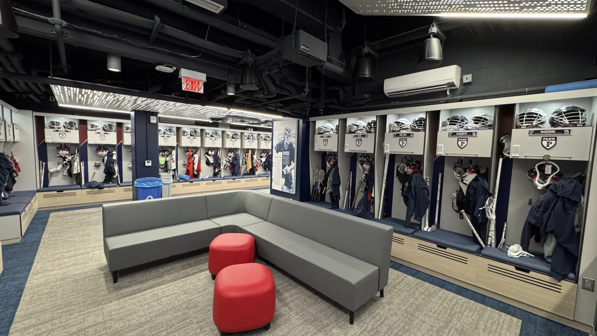 Modern sports team locker room with gray seating, red ottomans, hockey gear, and illuminated ceiling panels.