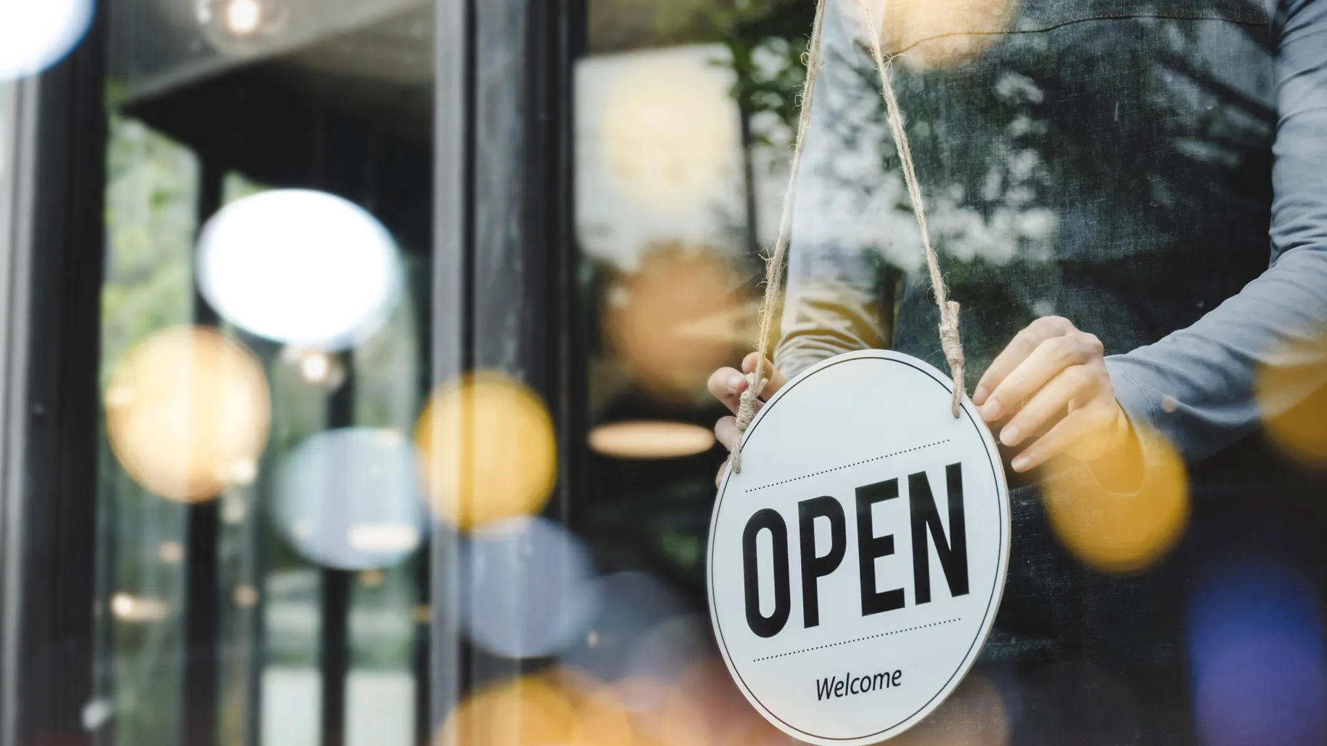 Person holding round open sign with welcome text in front of store window with reflections and bokeh lights