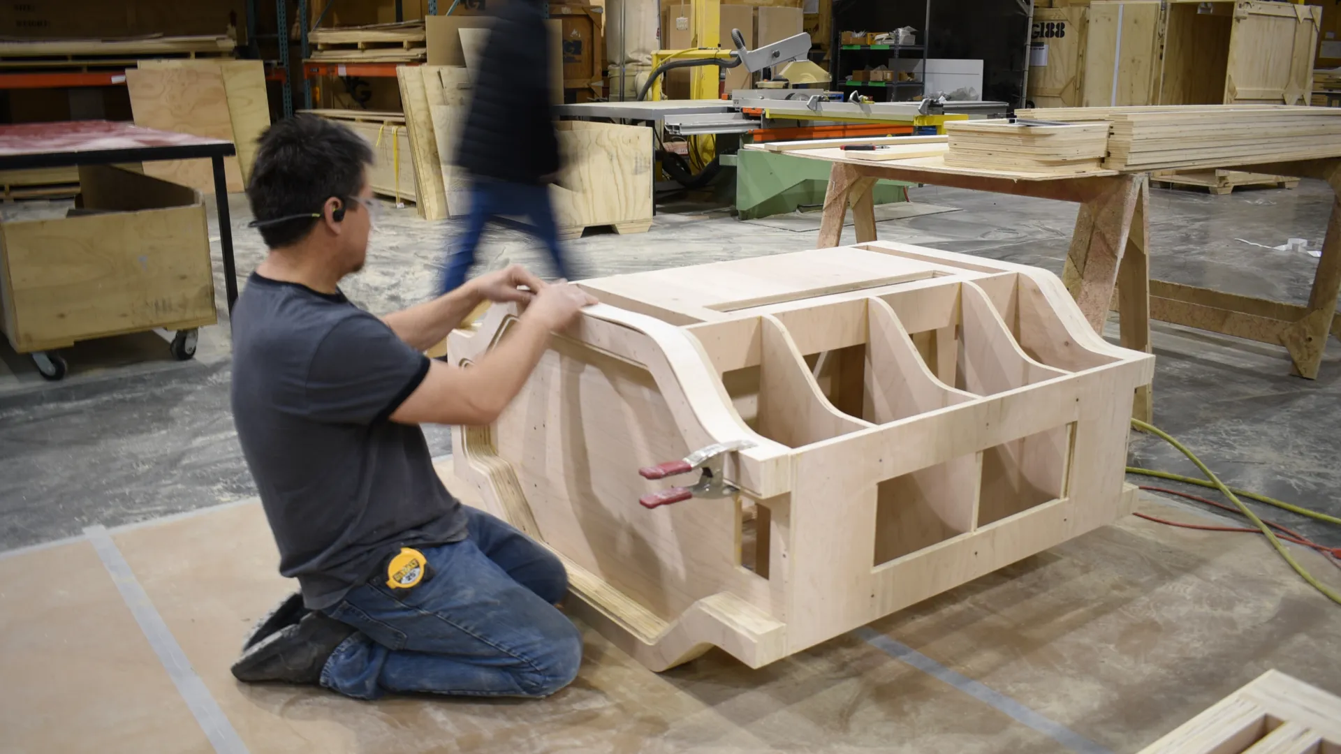Worker assembling a large wooden furniture piece in a busy workshop with tools and crates.