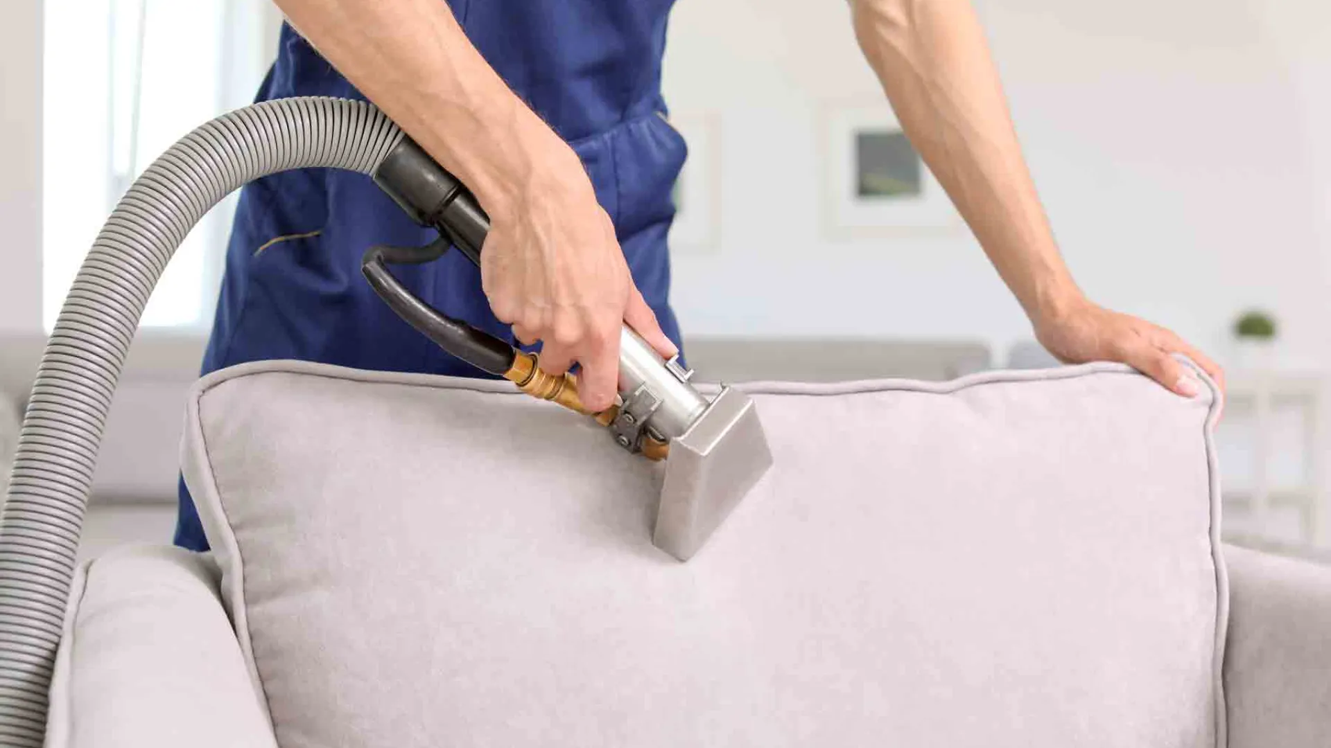 Person using a vacuum cleaner to clean a light gray sofa cushion in a bright room.