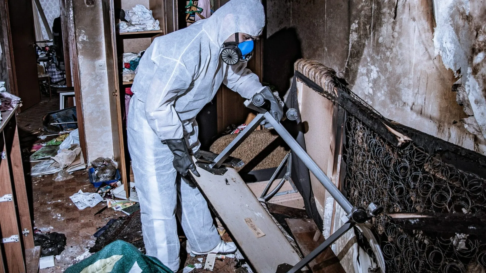 Person in protective suit and mask removing damaged furniture from a fire-damaged room with debris and soot