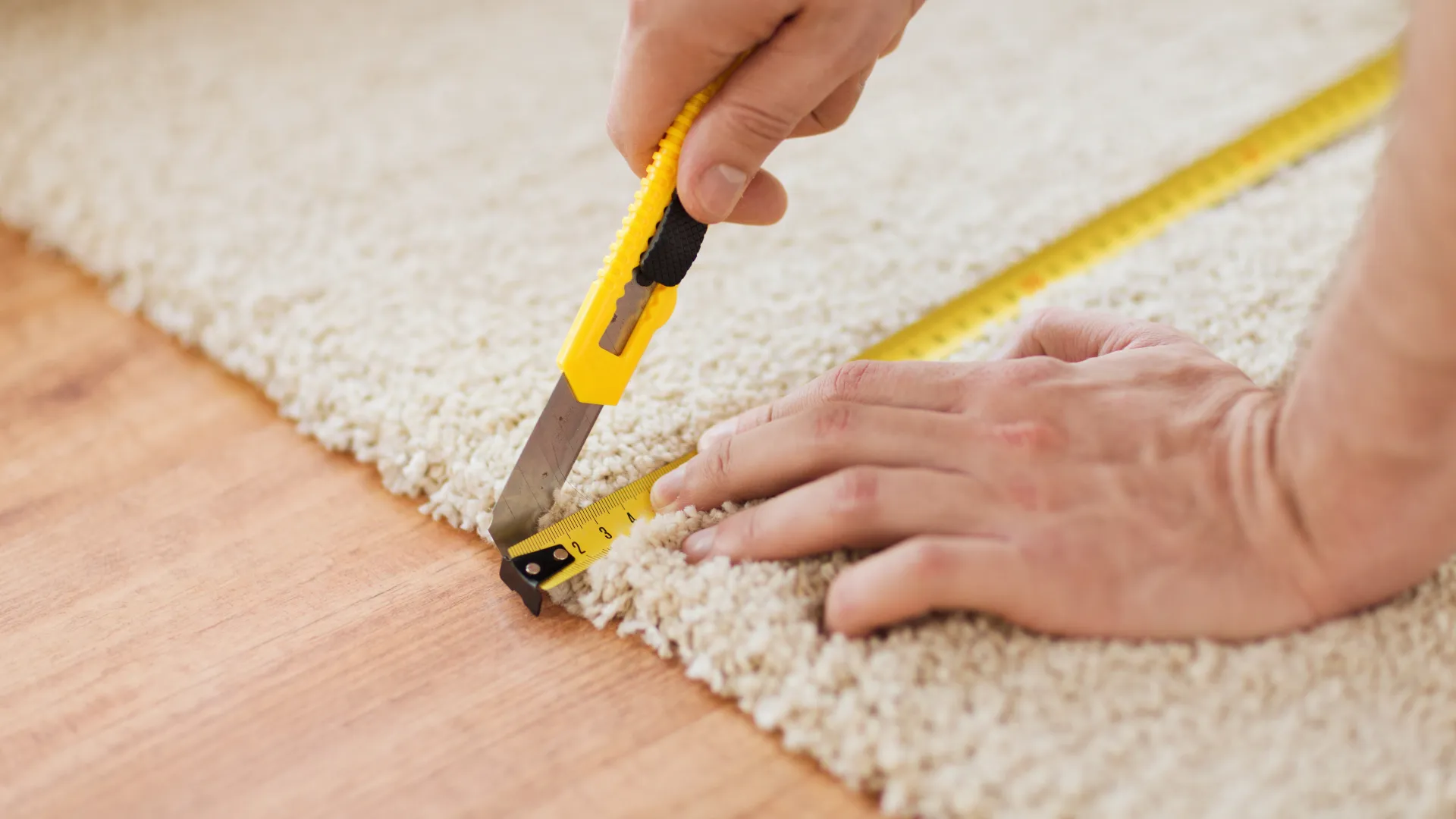 Person cutting beige carpet precisely along a measuring tape on wooden floor with a yellow utility knife.