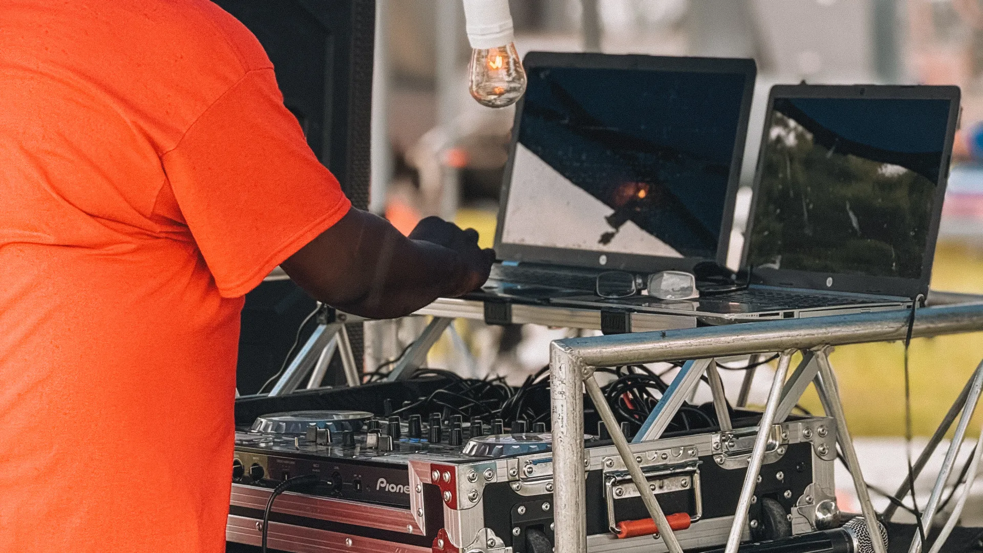 DJ in orange shirt operating mixing equipment under blue canopy at outdoor event with laptops and lights