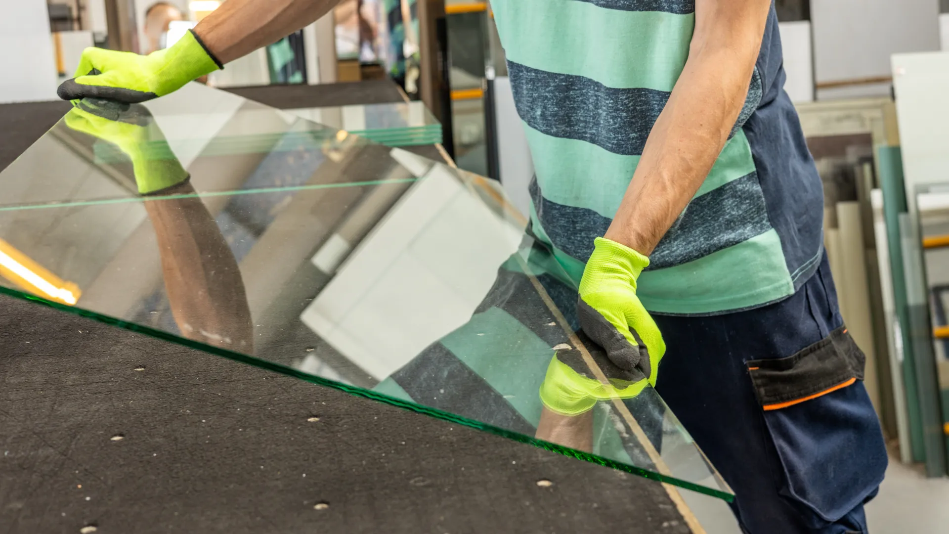 Worker wearing gloves handling large glass sheet in a workshop or factory setting.