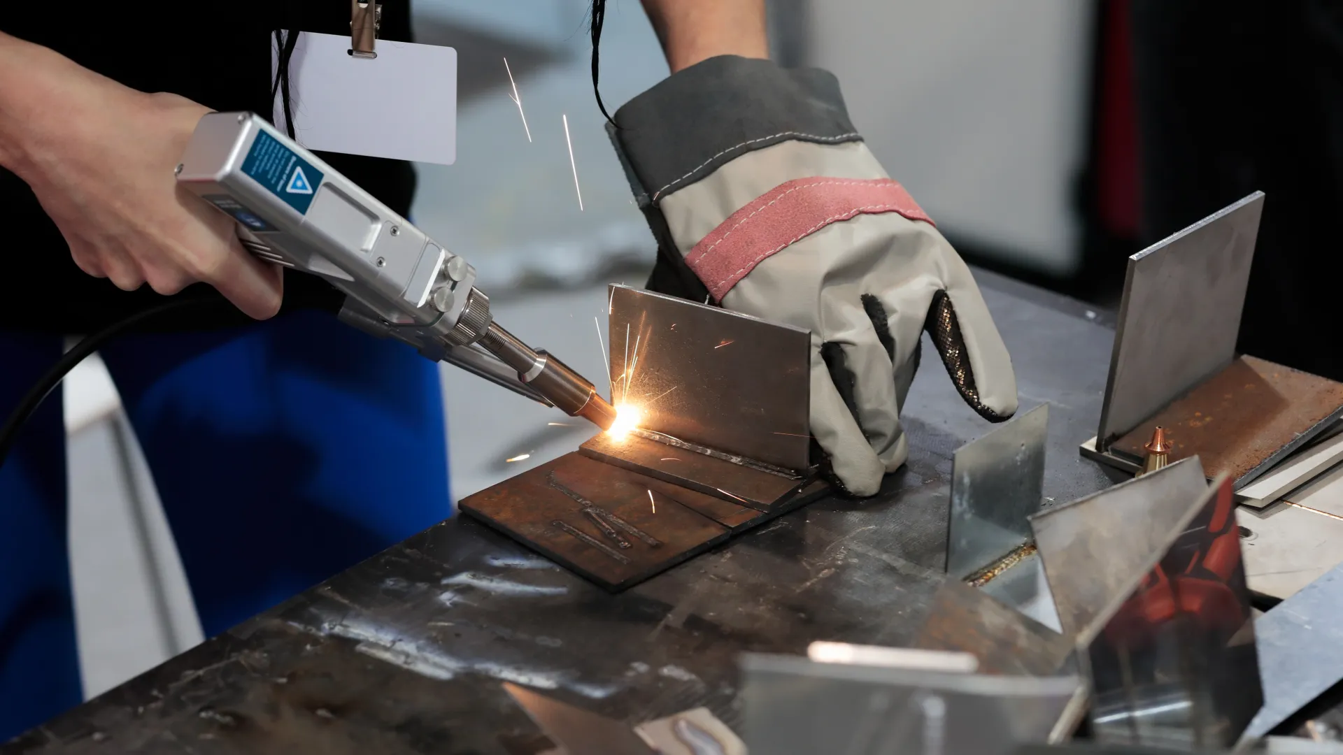 Close-up of a person welding metal pieces with sparks flying and wearing protective gloves and ID badge.