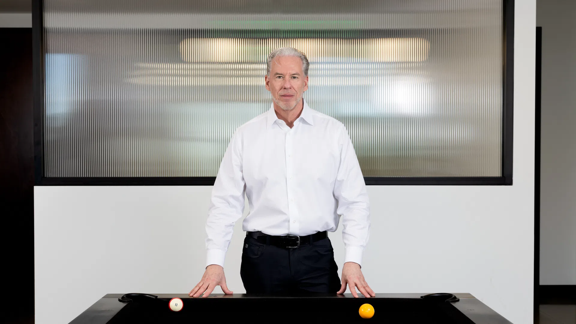 Mature man in white shirt standing behind a black pool table with scattered billiard balls indoors.