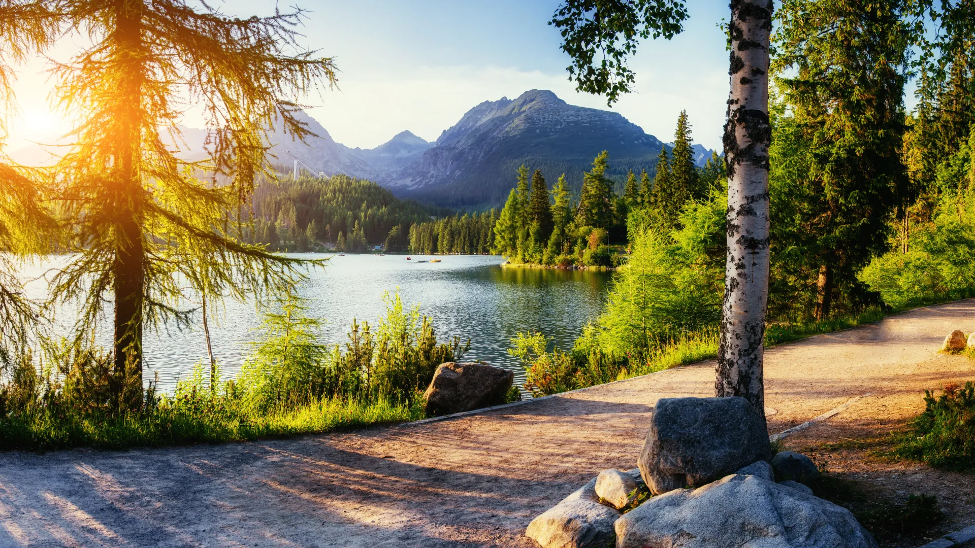 Sunlit lakeside path with trees, rocks, and mountains in the background on a clear day