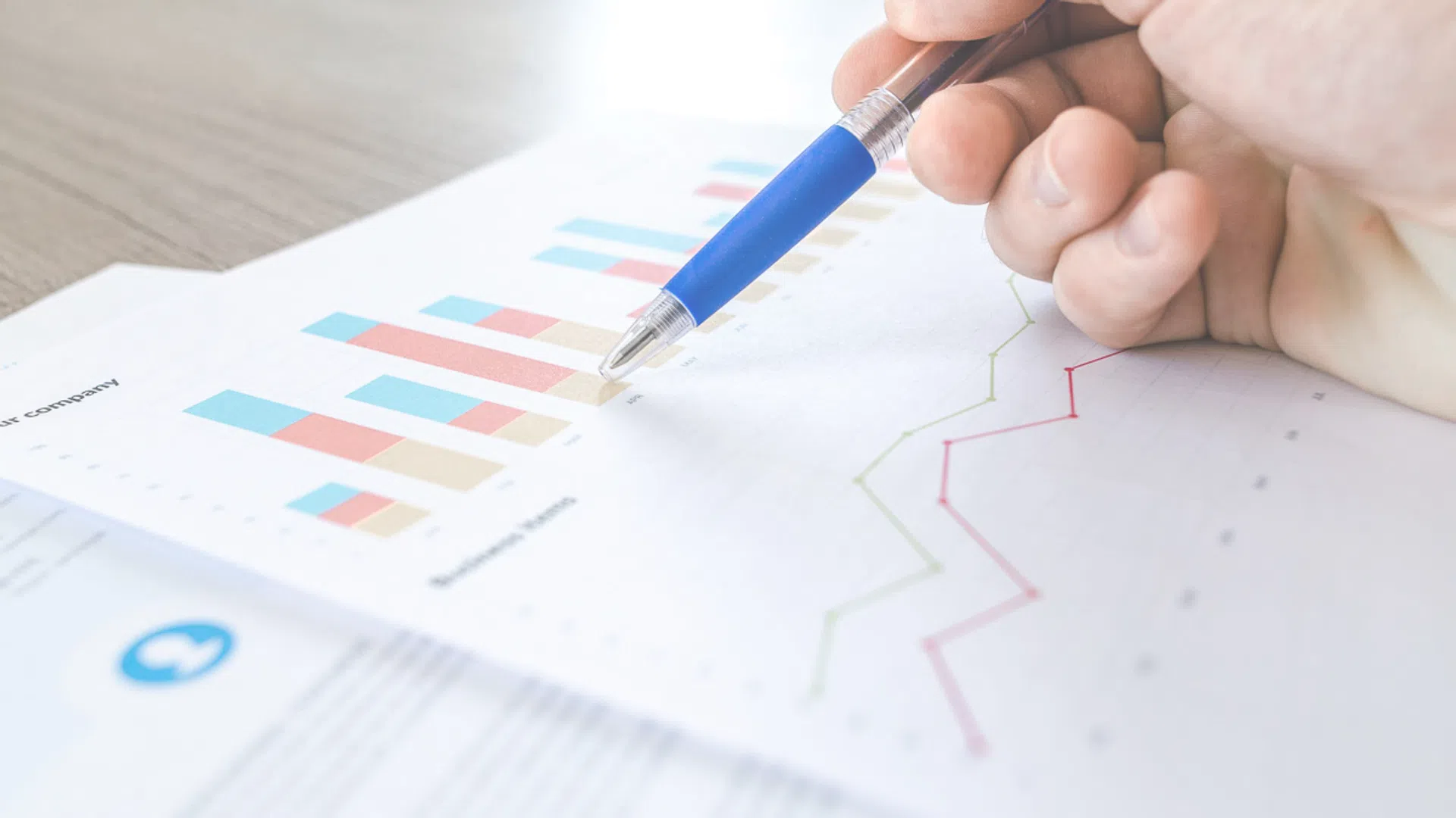 Hand holding a blue pen pointing to colorful bar and line charts on printed business reports on a wooden desk.