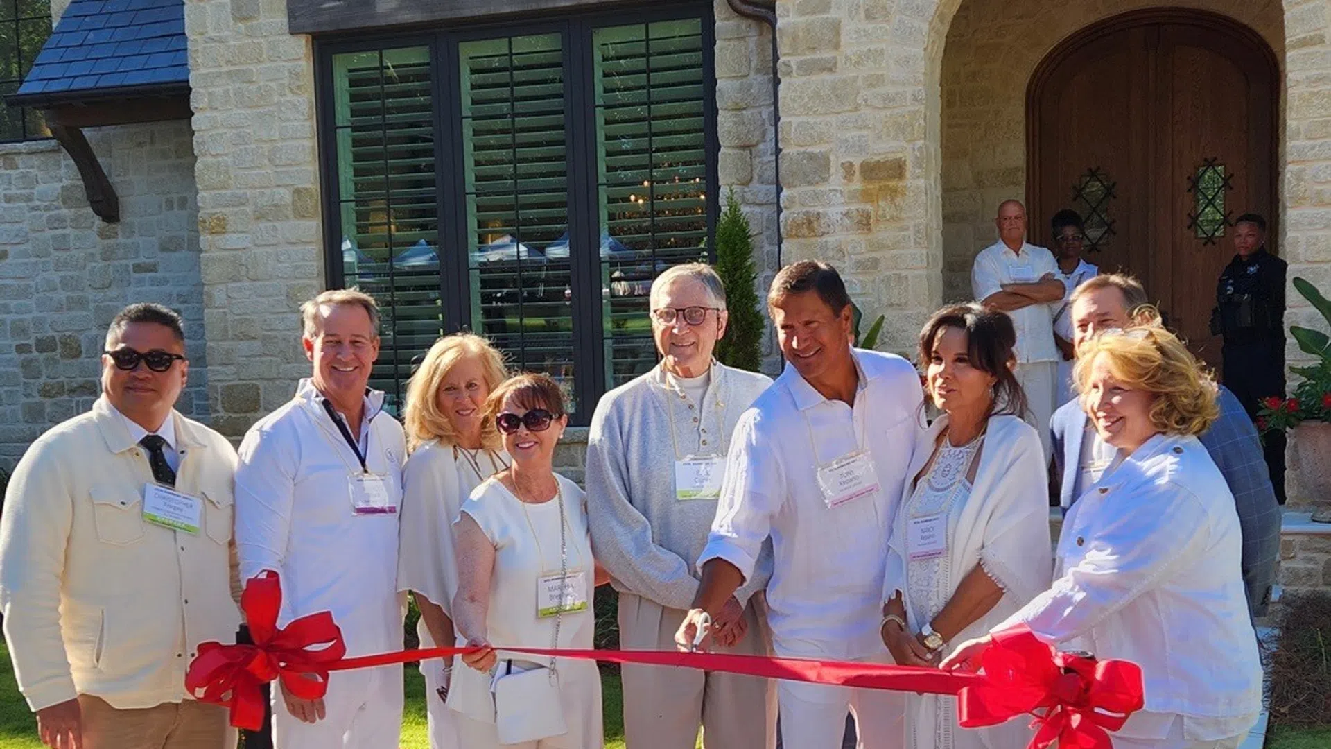 Group of people in white attire cutting a red ribbon at a house entrance during a sunny day event.