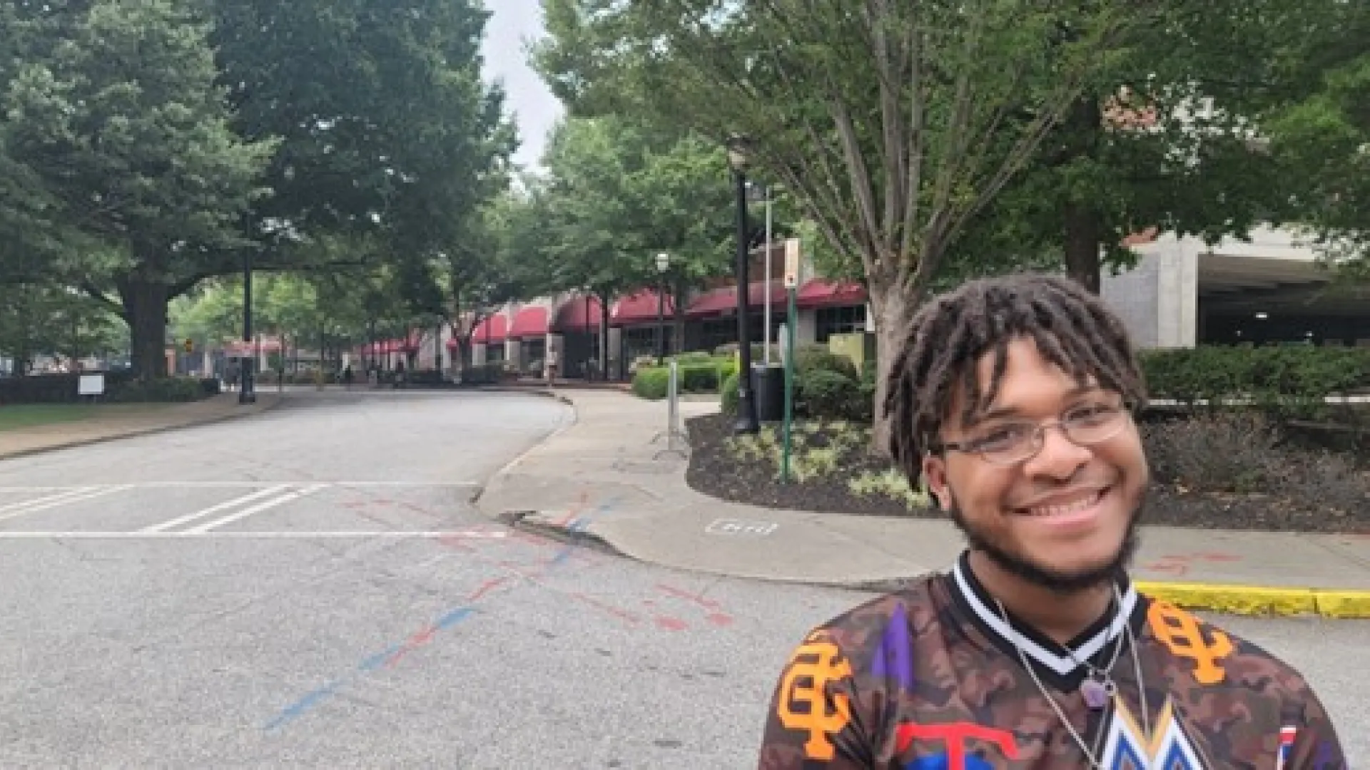 Smiling young man with glasses and dreadlocks stands on a quiet tree-lined street with red awnings in the background