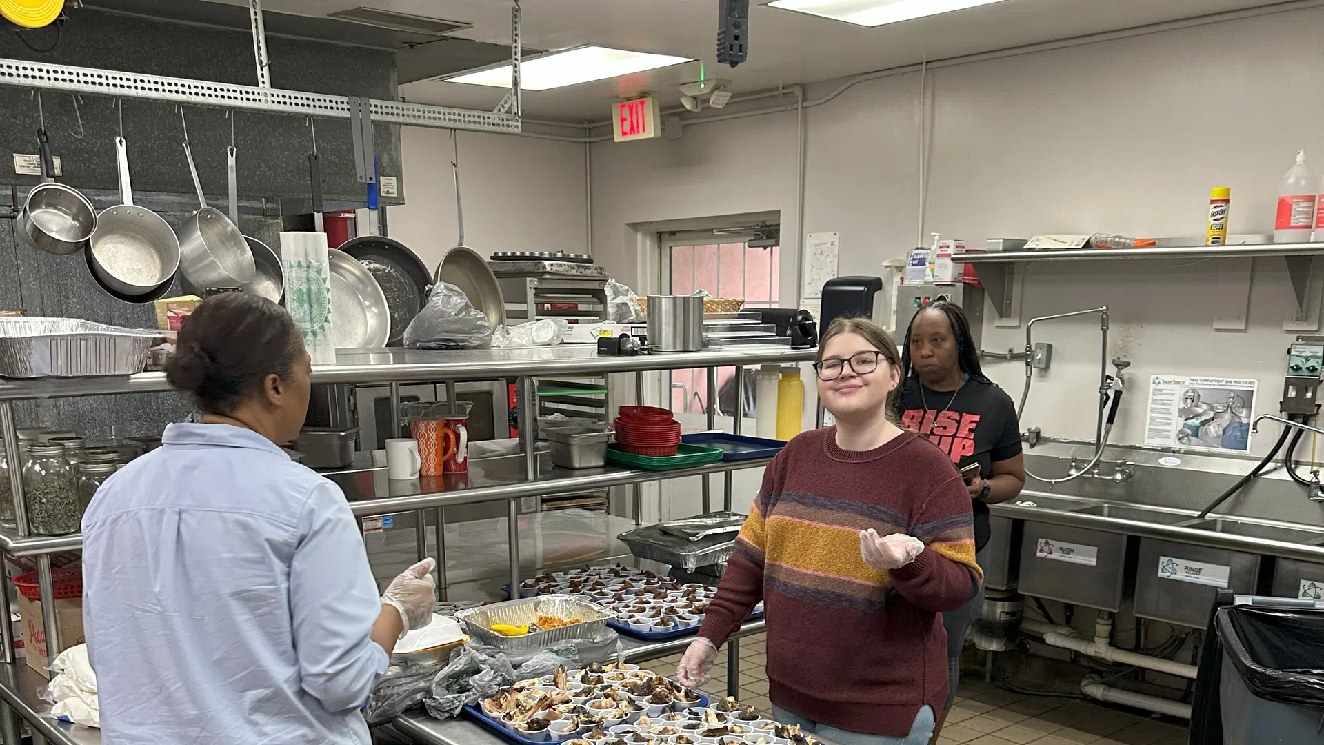 Three people preparing and arranging multiple trays of food samples in a commercial kitchen setting.