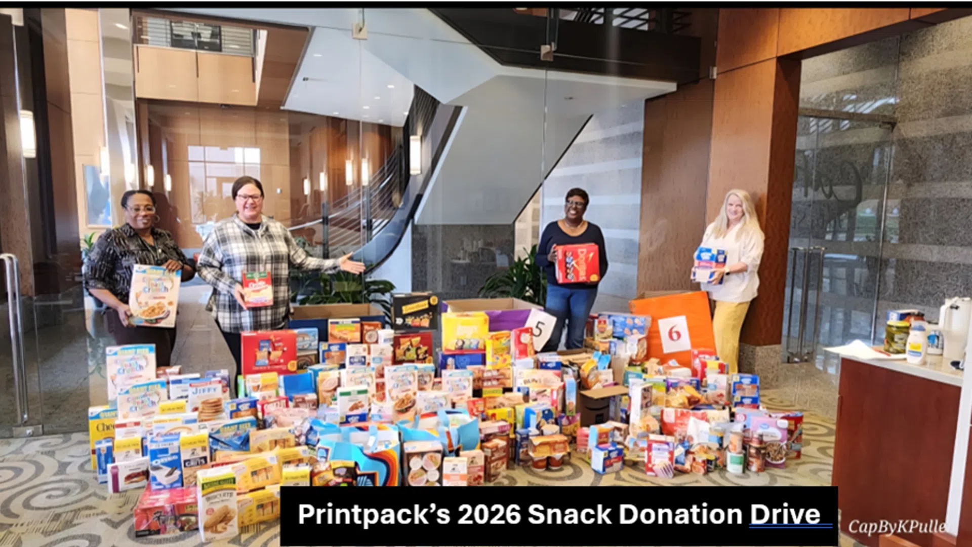 Four people stand behind a large collection of donated snacks in an office lobby for Printpack's 2026 charity drive.